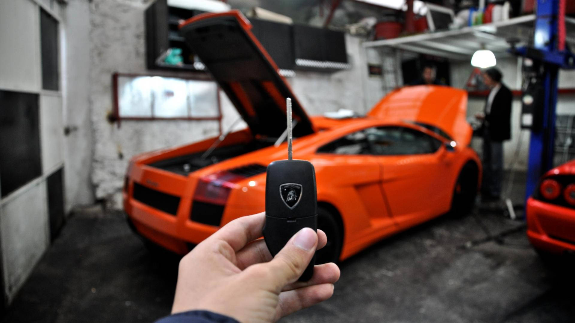 橘色 桌布 壁紙 — 一隻手在雜亂的車庫中，在引擎蓋打開的亮橙色藍寶堅尼前面，拿著一個帶有銀色鑰匙葉的黑色車鑰匙遙控器。
