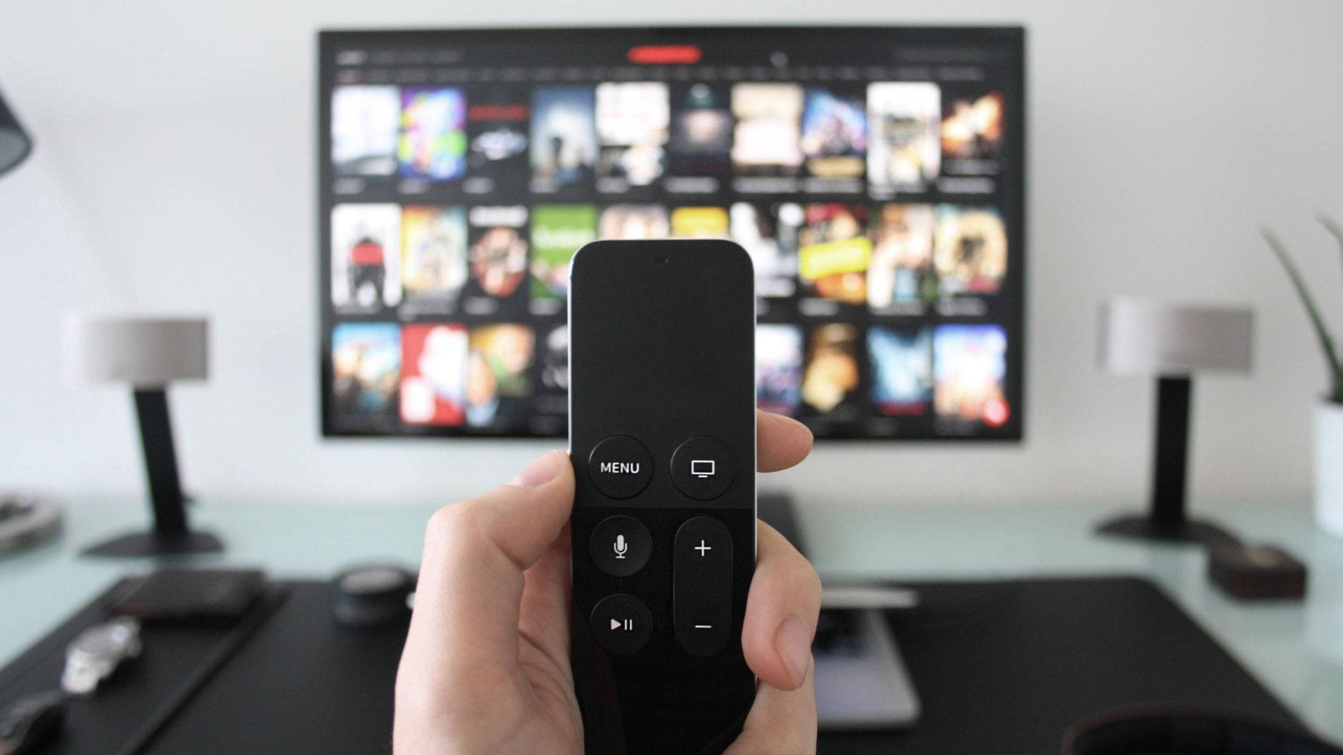 tv 桌布 壁紙 — 一隻手在電視前面拿著一個黑色遙控器，電視顯示一個有彩色縮圖的串流服務介面。