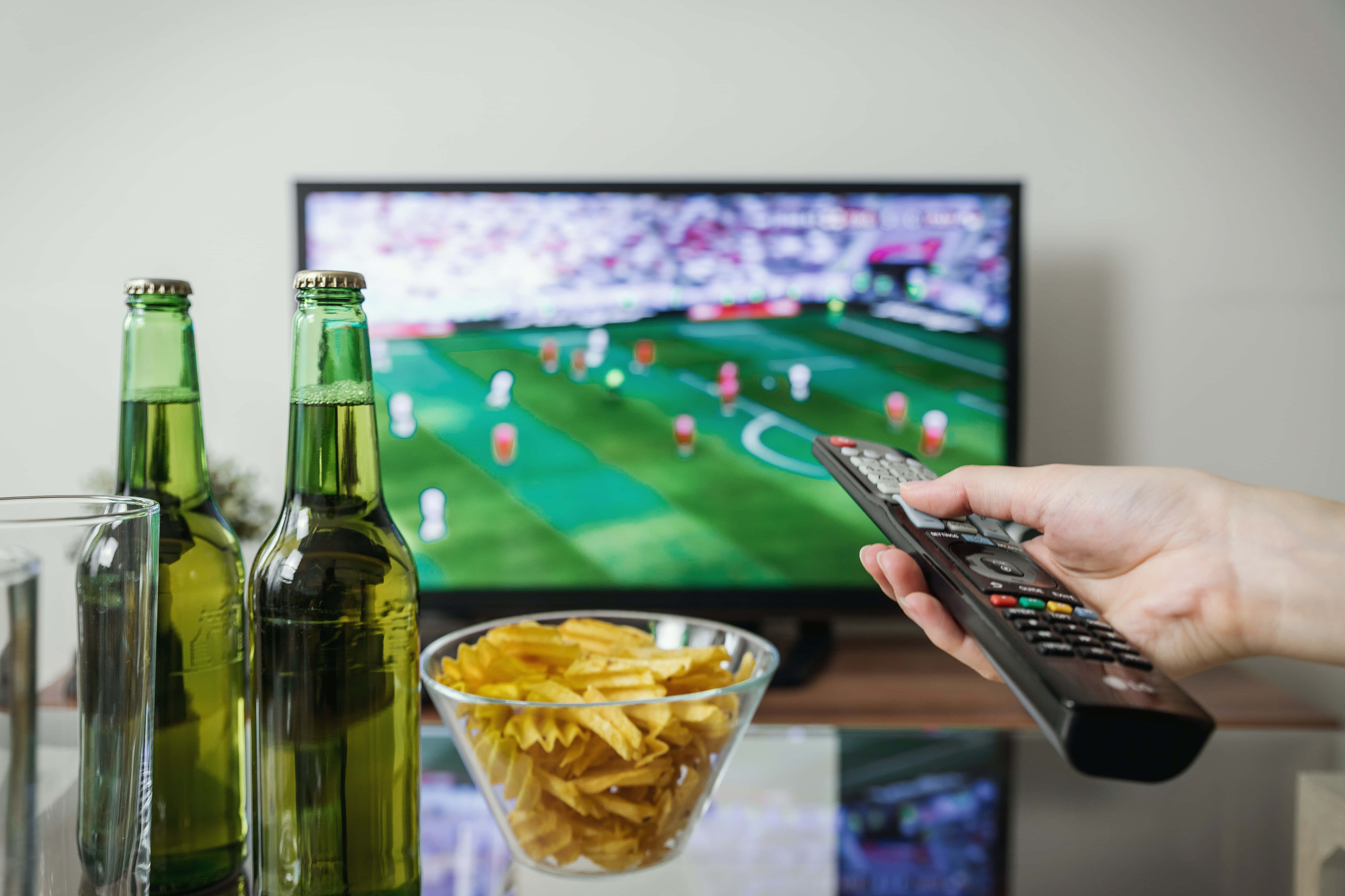 television 桌布 壁紙 — 一隻手在電視前面拿著遙控器，電視正在播放足球比賽，桌上有綠色啤酒瓶和一碗薯片。