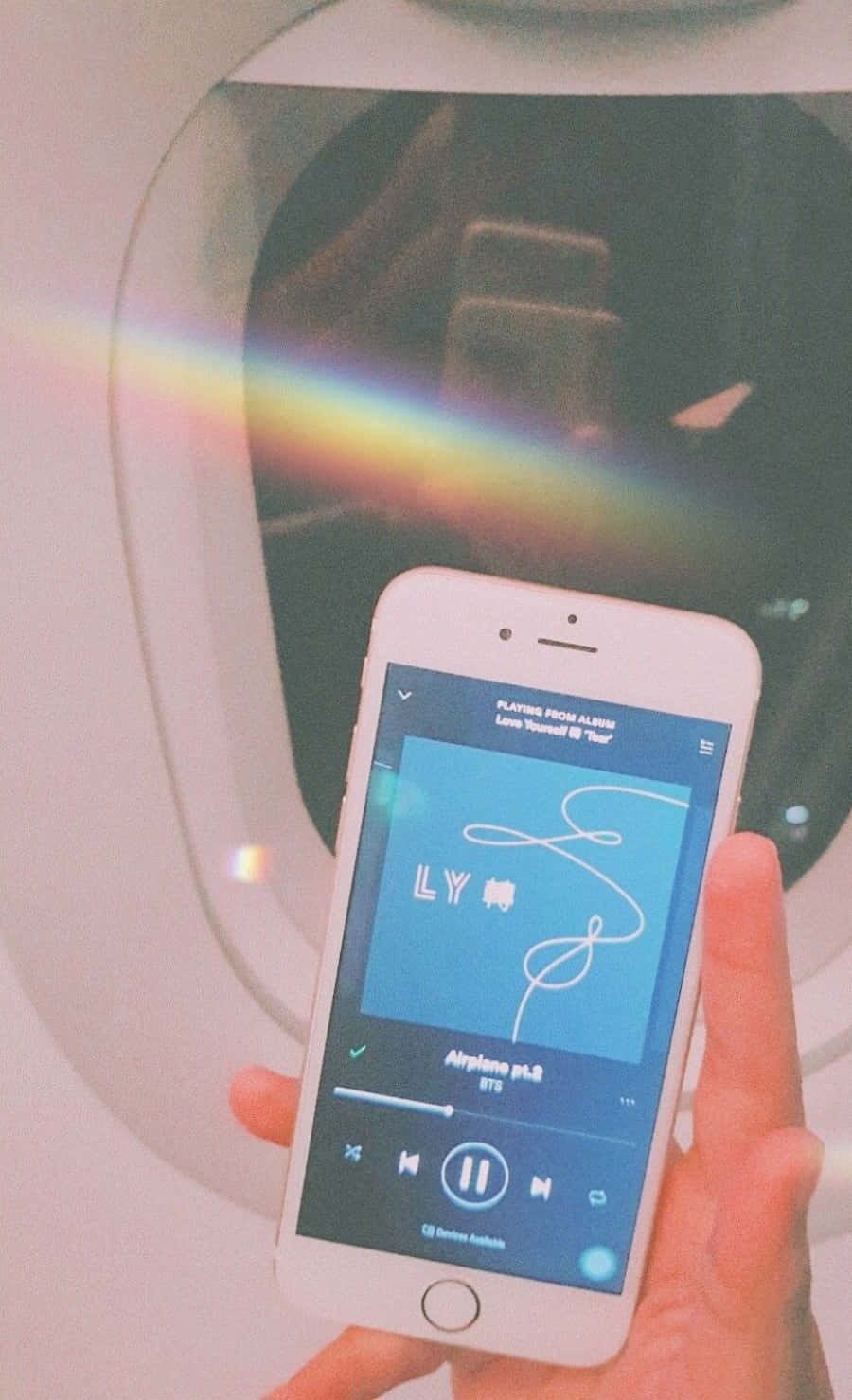 手機 桌布 壁紙 — 一隻手在飛機窗附近拿著手機播放BTS的《Airplane pt.2》，窗上有彩虹稜鏡效果。