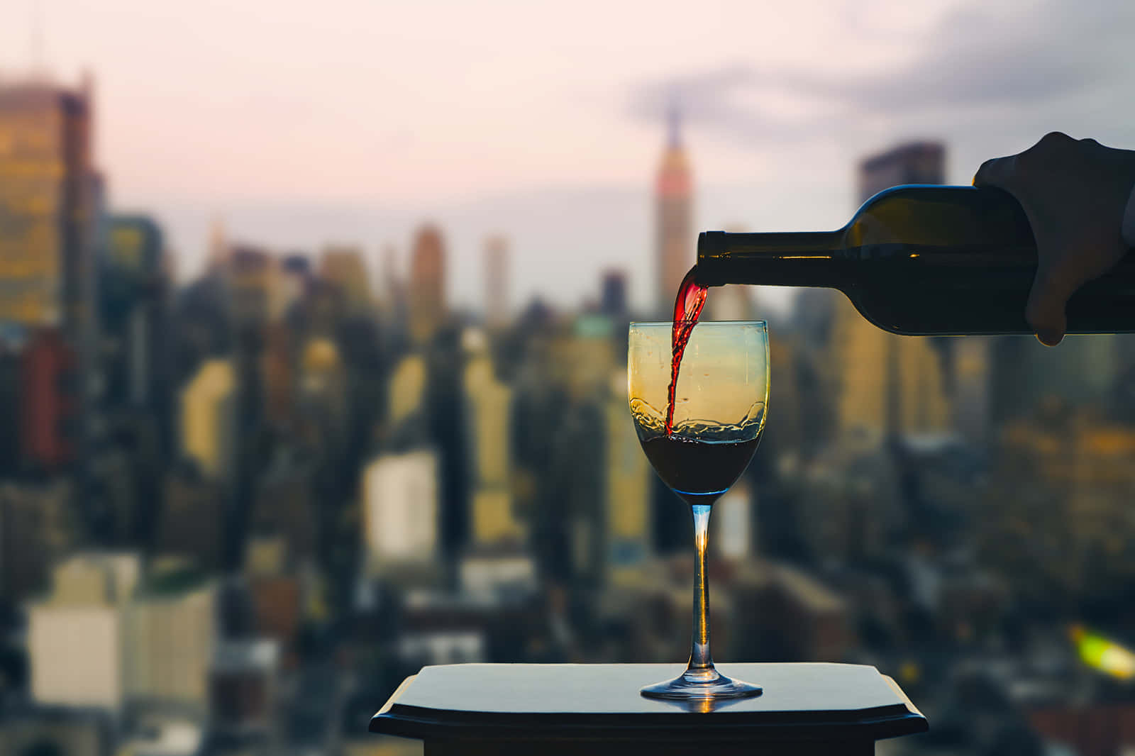 Nyc 桌布 壁紙 — 一隻手在黃昏時分將紅酒倒入玻璃杯，背景是模糊的城市天際線。