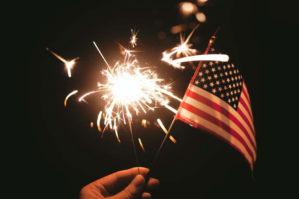 Happy Fourth Of July 桌布 壁紙 — 一隻手在黑暗夜空背景下，在小美國國旗旁邊拿著一支明亮的火花煙火。