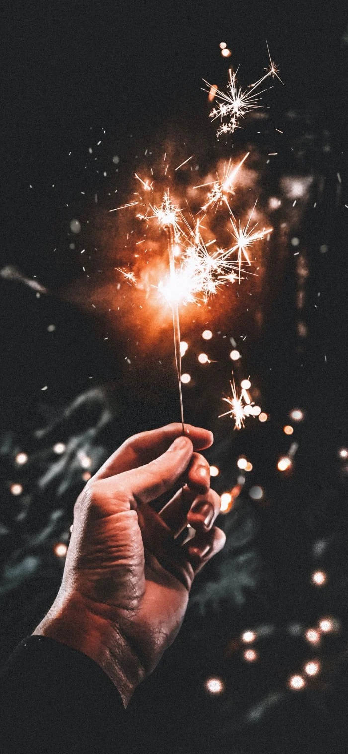 Fireworks 桌布 壁紙 — 一隻手在黑暗的夜空下拿著發光的煙火棒，明亮的橙色火花向外飛散。
