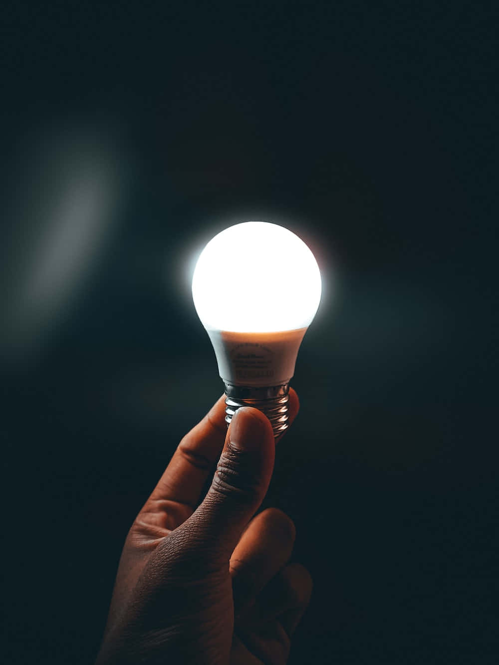 Light 桌布 壁紙 — 一隻手在黑暗背景下拿著發光的燈泡，在手指上投射出柔和的光線。