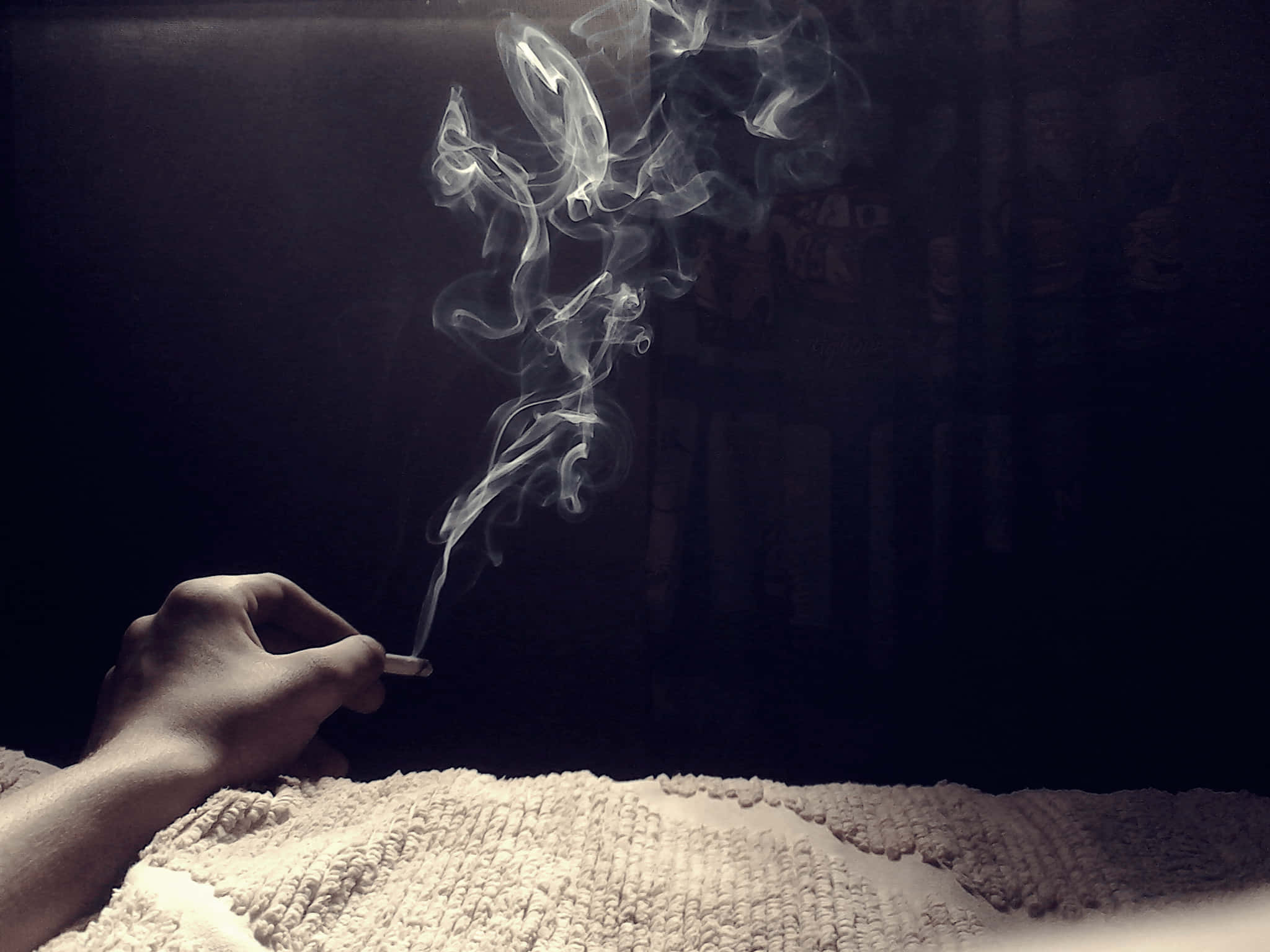 cigarettes 桌布 壁紙 — 一隻手在黑暗背景下拿著點燃的香煙，旋轉的煙霧在帶有紋理的白色表面上升起。