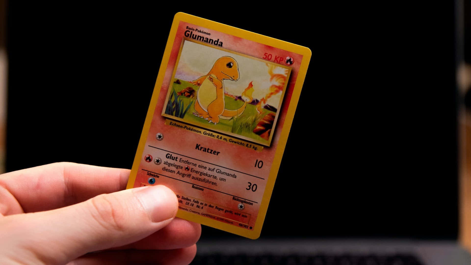 寶可夢 桌布 壁紙 — 一隻手在黑暗背景中拿著一張德文Glumanda Pokémon卡，背景可見一部筆記型電腦。
