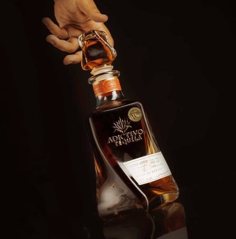 電腦 桌布 壁紙 — 一隻手在黑暗背景前拿著一瓶Adictivo Tequila，戲劇性的燈光突顯了琥珀色的液體和標籤細節。