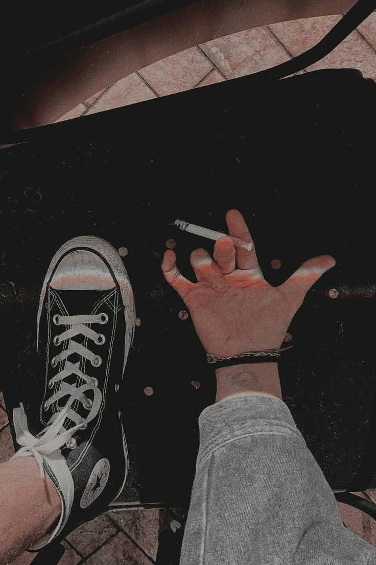垃圾美學 桌布 壁紙 — 一隻手在黑色Converse運動鞋旁邊拿著一支煙，背景是深色表面和瓷磚地板。