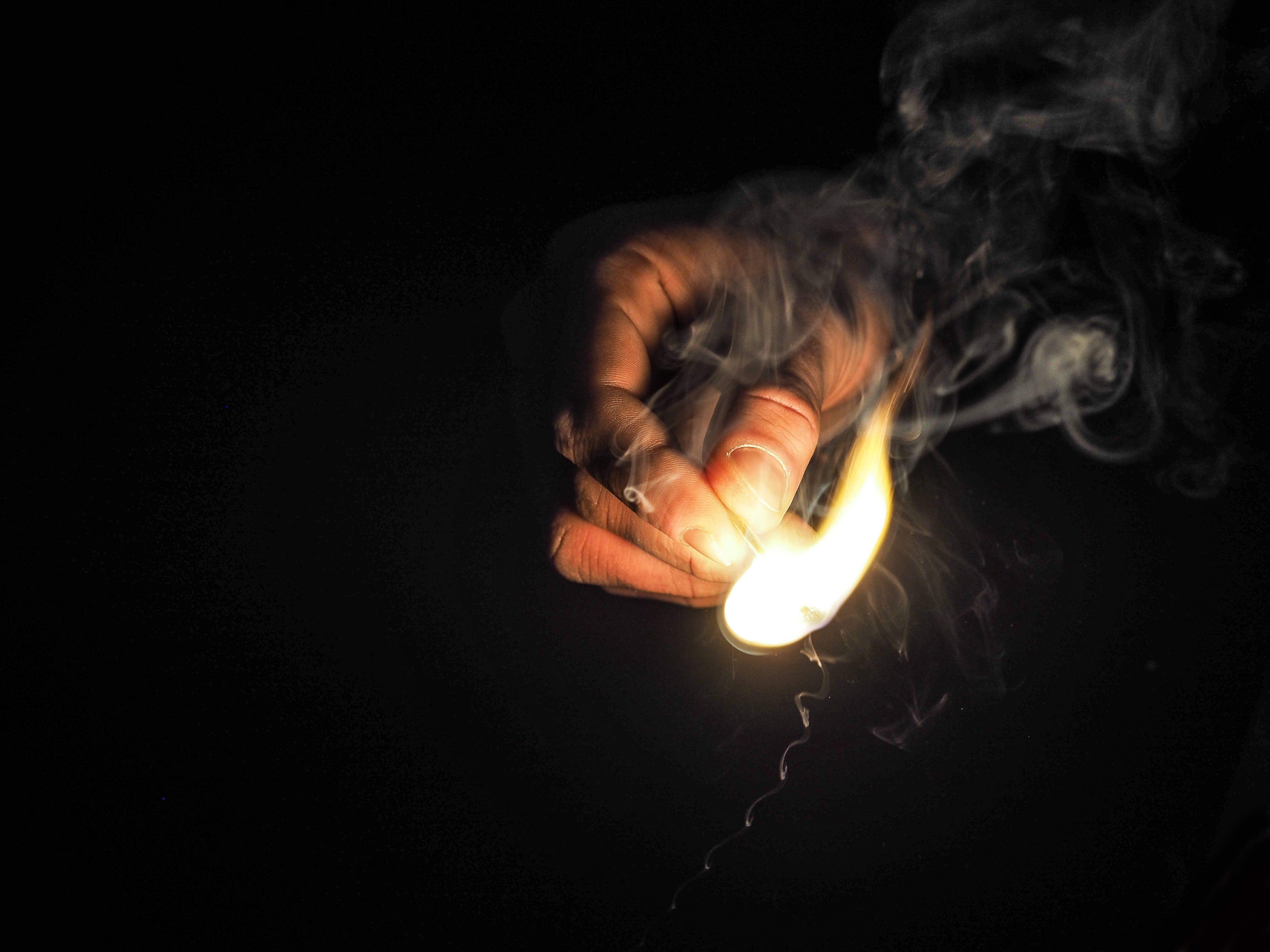 火焰動畫 桌布 壁紙 — 一隻手在黑色背景前拿著一根發光的火柴，火焰中升起縷縷煙霧。
