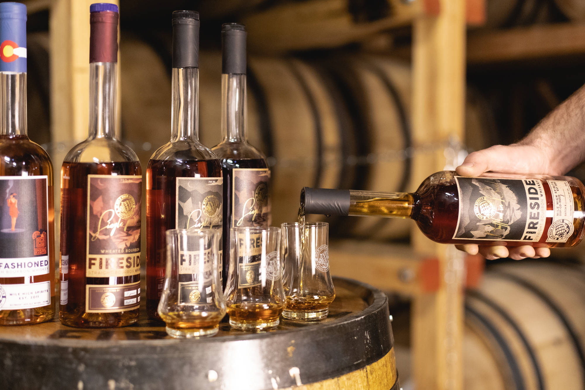 alcohol 桌布 壁紙 — 一隻手將Fireside酒瓶中的琥珀色威士忌倒入放在木桶上的品酒杯中，背景是溫暖燈光的釀酒廠。