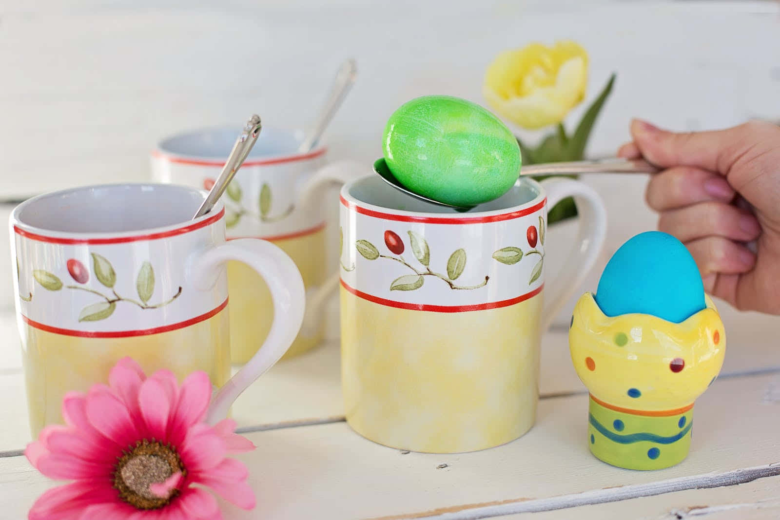 Pastel Easter 桌布 壁紙 — 一隻手將一個綠色雞蛋放入一個帶有葉子圖案的黃色杯子中，旁邊有一朵粉紅色的花和一個藍色的雞蛋放在白色木質表面上。