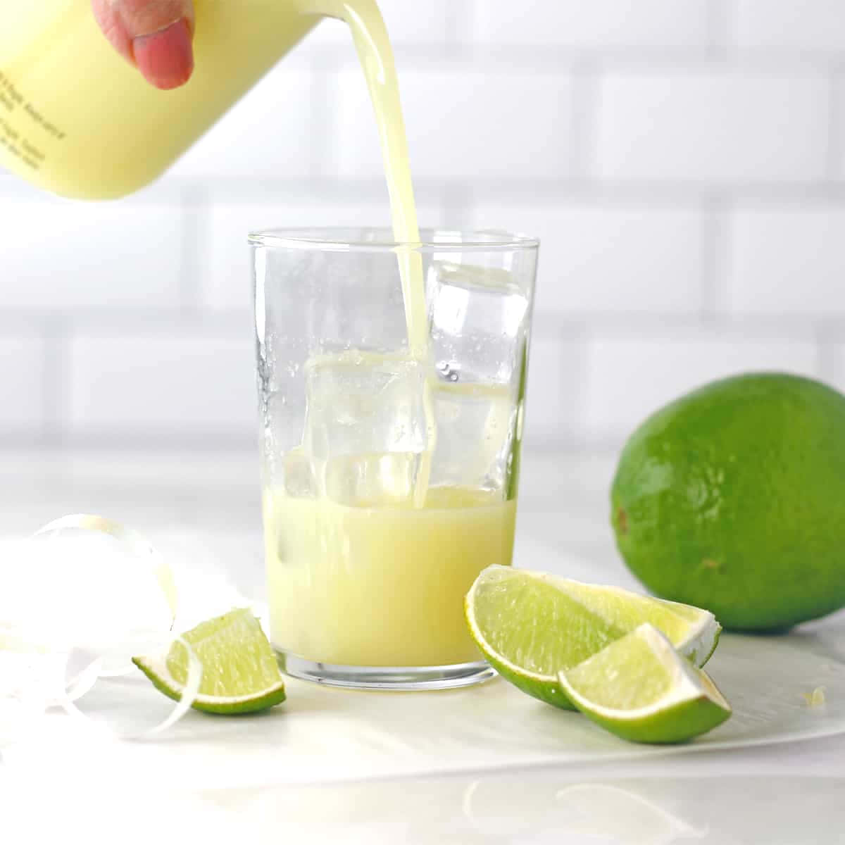 Lime 桌布 壁紙 — 一隻手將充滿活力的黃色萊姆水倒在冰上，倒入玻璃杯中，旁邊有新鮮萊姆角和一個完整的萊姆，放在白色瓷磚檯面上。