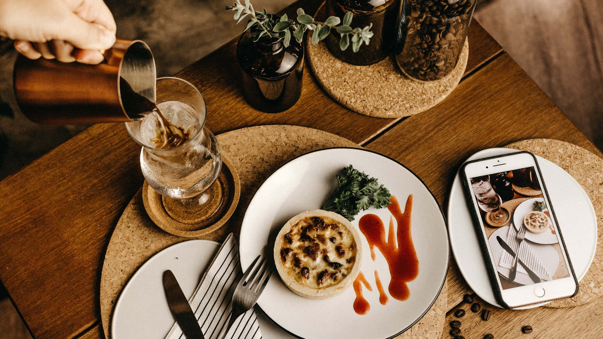Food Table 桌布 壁紙 — 一隻手將咖啡倒入玻璃杯，旁邊有一碟法式鹹派和一部顯示相同餐點的手機，放在木桌上。