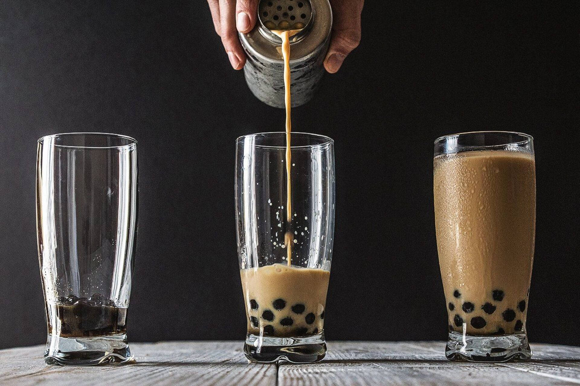 Bubble 桌布 壁紙 — 一隻手將奶油泡沫茶倒入裝滿黑色木薯珍珠的玻璃杯中，背景為深色。