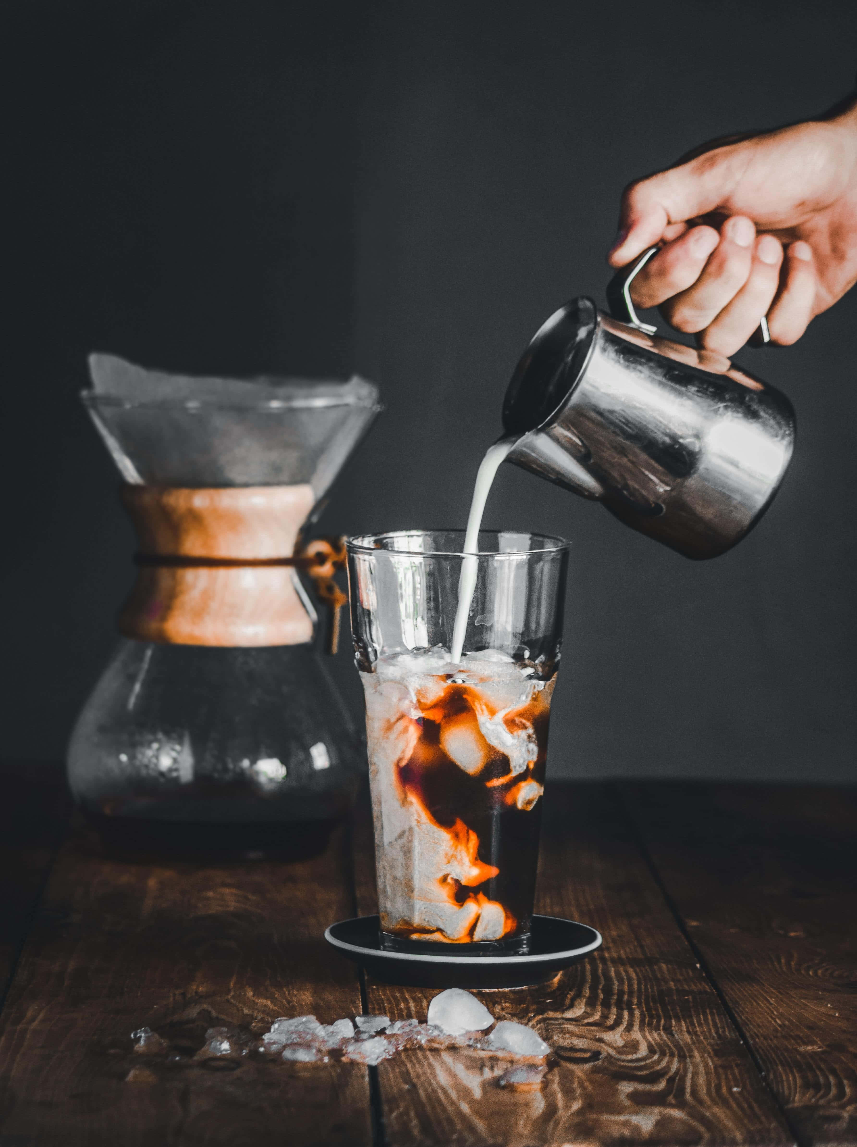 Iced Coffee 桌布 壁紙 — 一隻手將奶油牛奶倒入一杯加冰的冰咖啡中，放在深色木桌上。