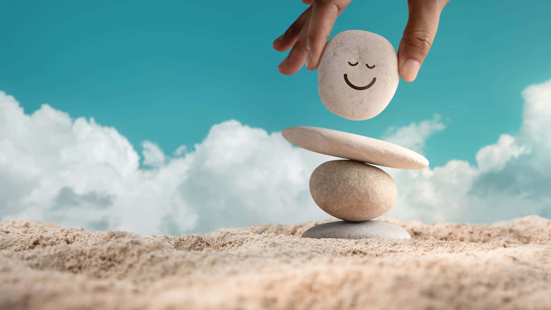 Smiley 桌布 壁紙 — 一隻手將微笑的石頭放在沙灘上平衡的石堆上，藍天白雲下。