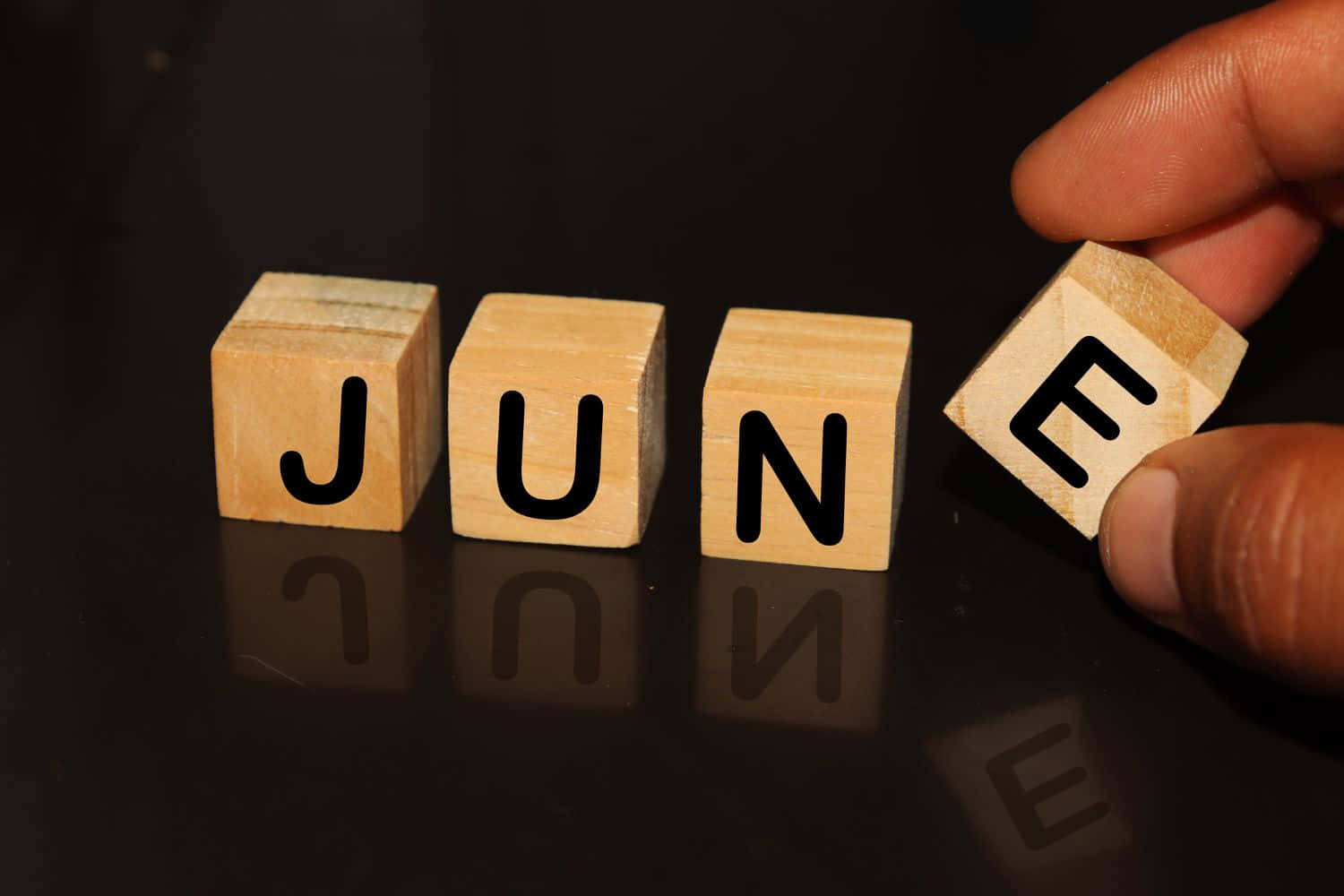 June 桌布 壁紙 — 一隻手將拼出JUNE的木塊放在帶有柔和光線的反射黑色表面上。