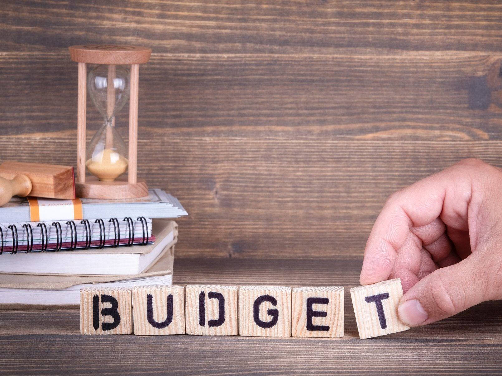 Word 桌布 壁紙 — 一隻手將拼寫「BUDGET」的木塊放在堆疊的筆記本和沙漏旁邊的質樸木質表面上。