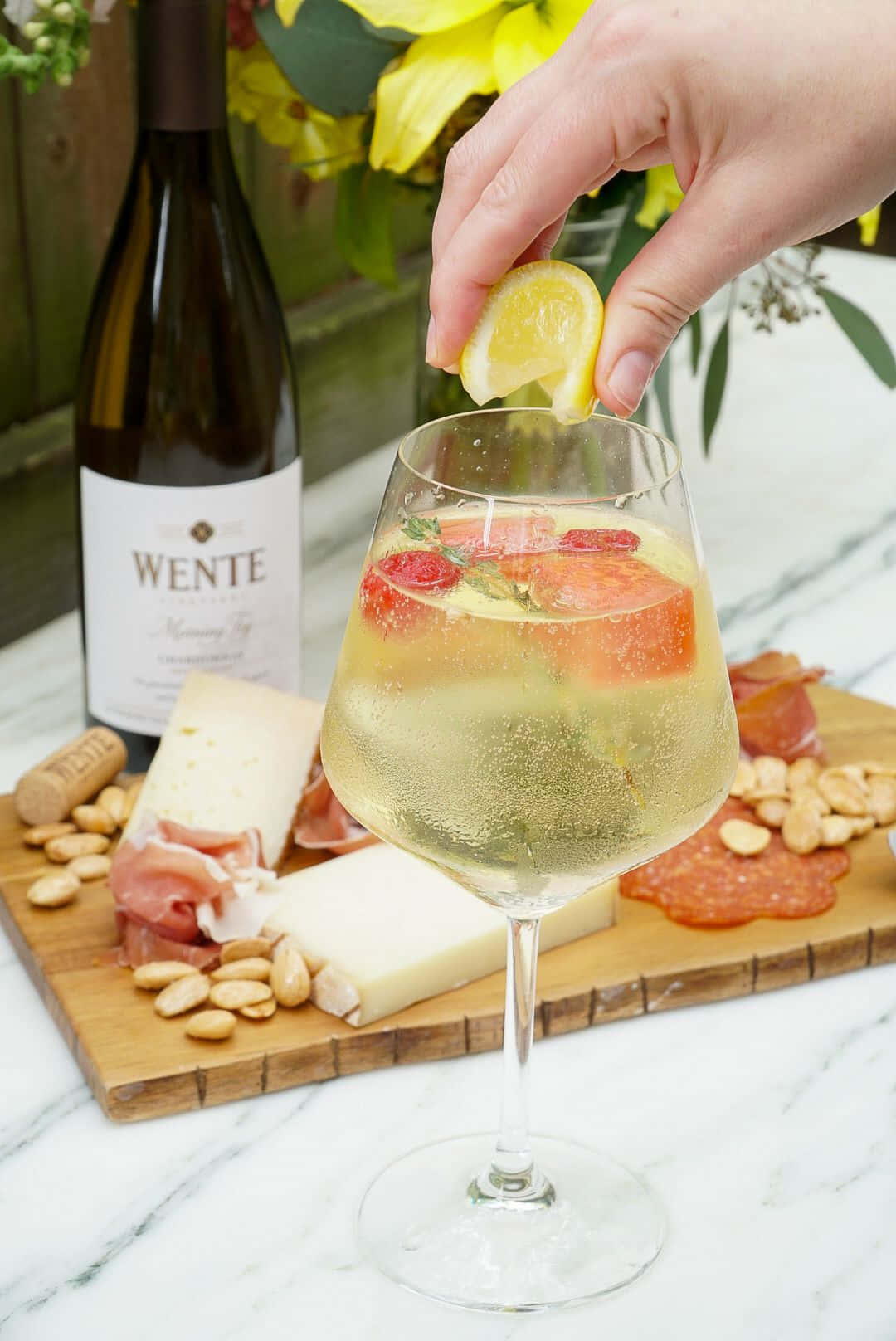 White Wine 桌布 壁紙 — 一隻手將檸檬角擠入一杯氣泡酒中，周圍有大理石檯面上的起司、帕爾馬火腿和堅果。