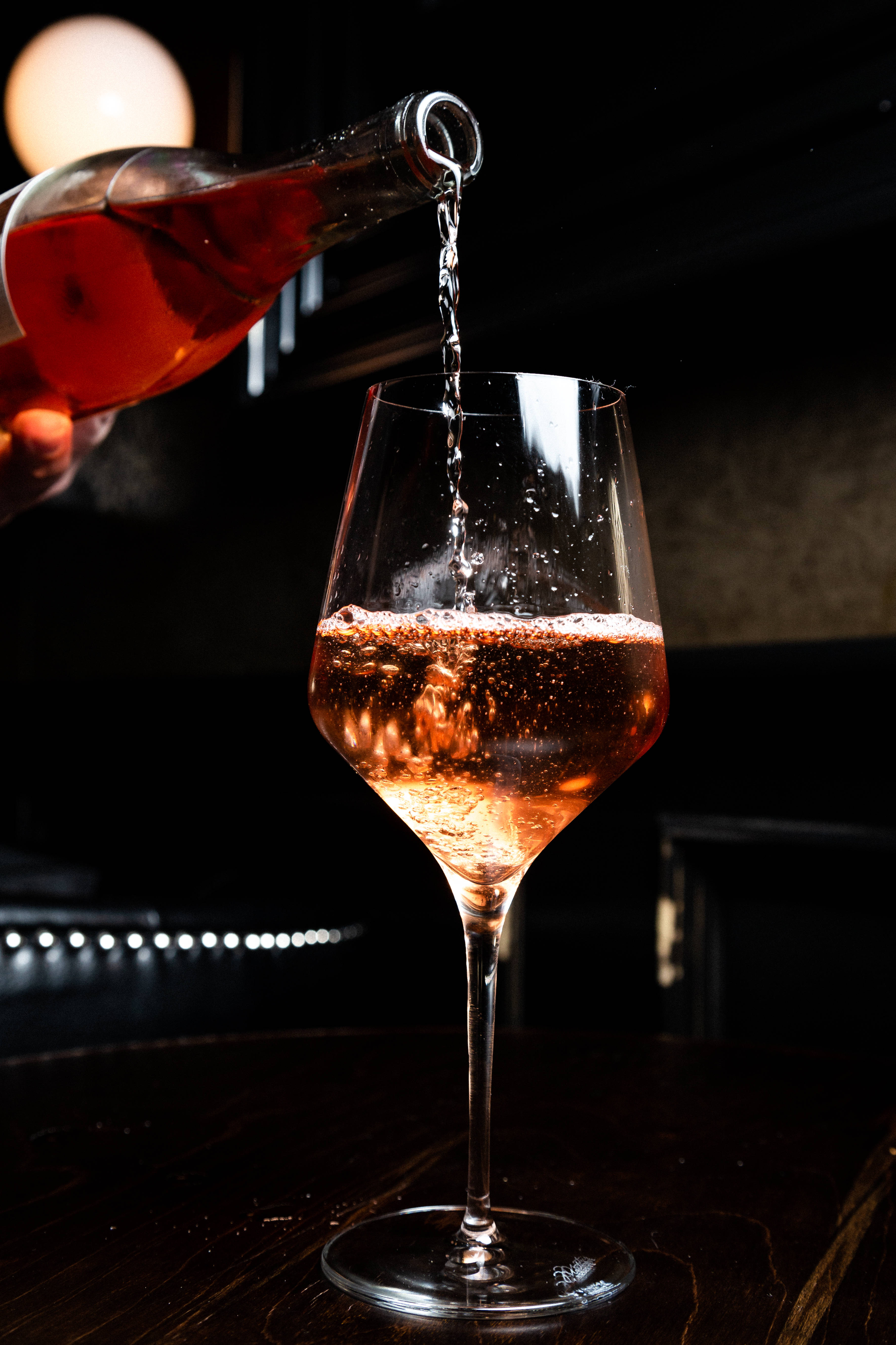 葡萄酒 桌布 壁紙 — 一隻手將氣泡玫瑰酒倒入玻璃杯中，背景是昏暗的酒吧和溫暖的環境燈光。