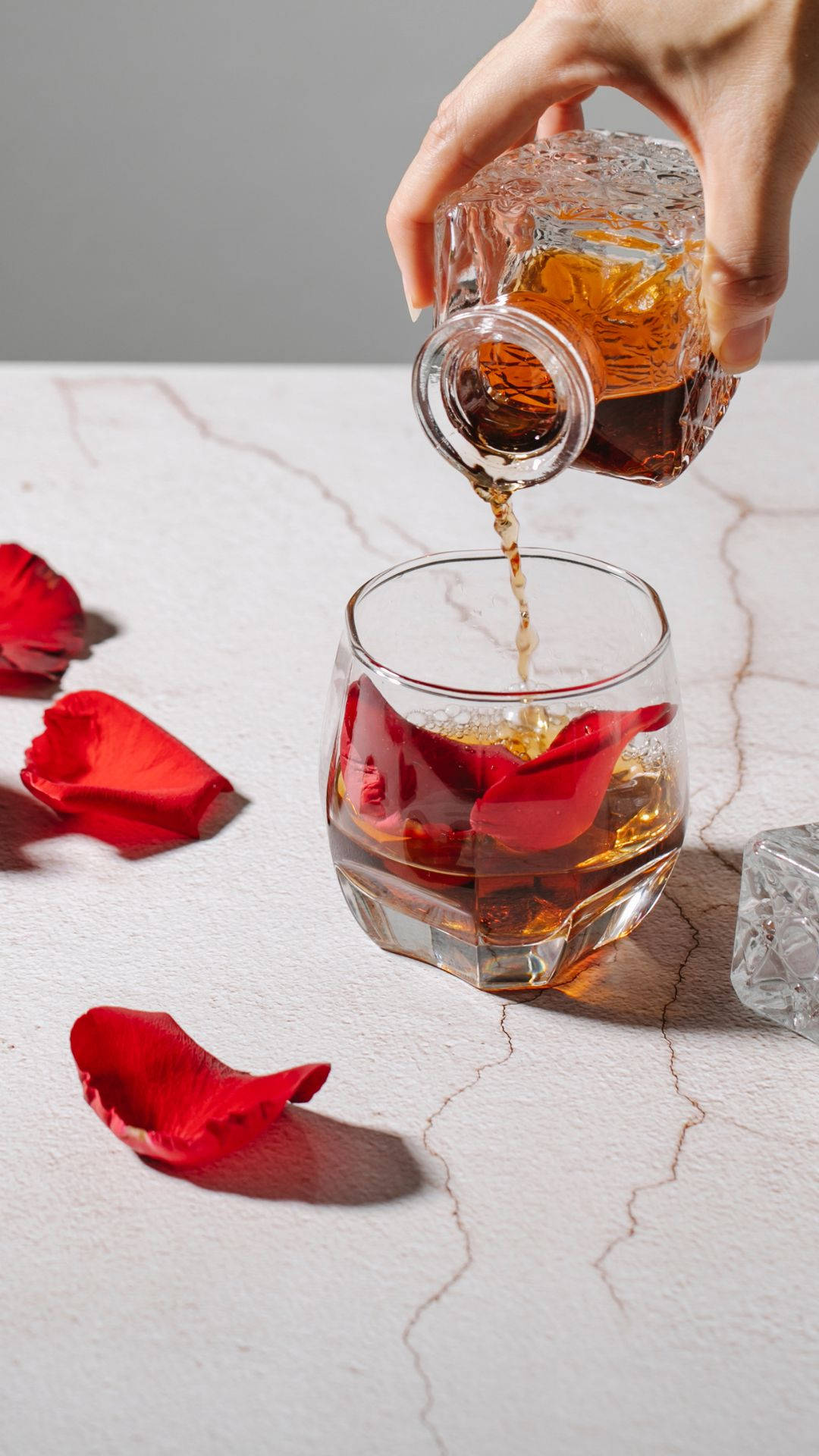 Drink 桌布 壁紙 — 一隻手將水晶瓶中的琥珀色液體倒入一個裝有紅玫瑰花瓣和冰塊的玻璃杯中，置於白色大理石表面上。
