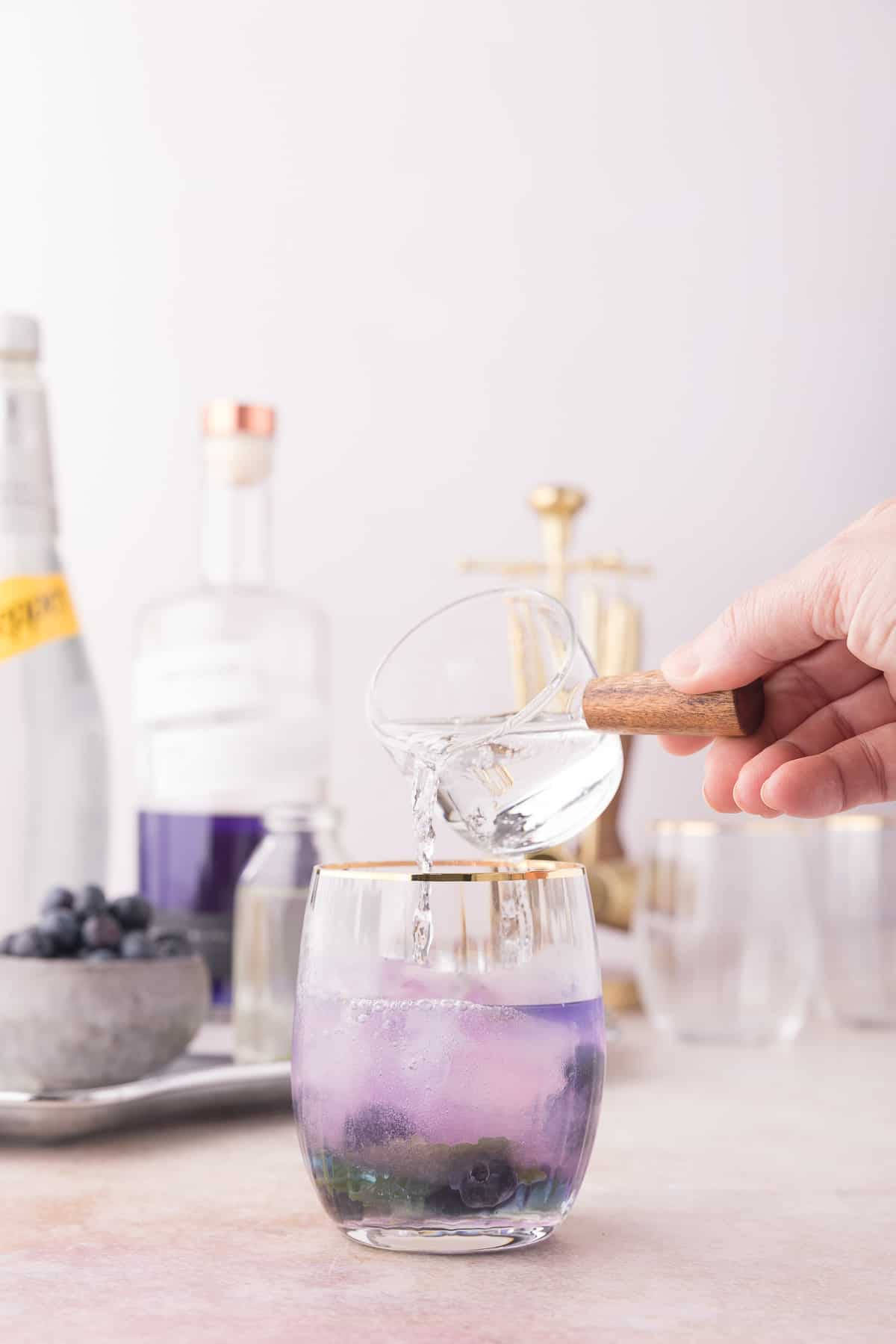 cocktail 桌布 壁紙 — 一隻手將清澈液體倒入一個帶有紫色藍莓飲料和金色邊緣的玻璃杯中，置於柔軟的粉紅色表面上。