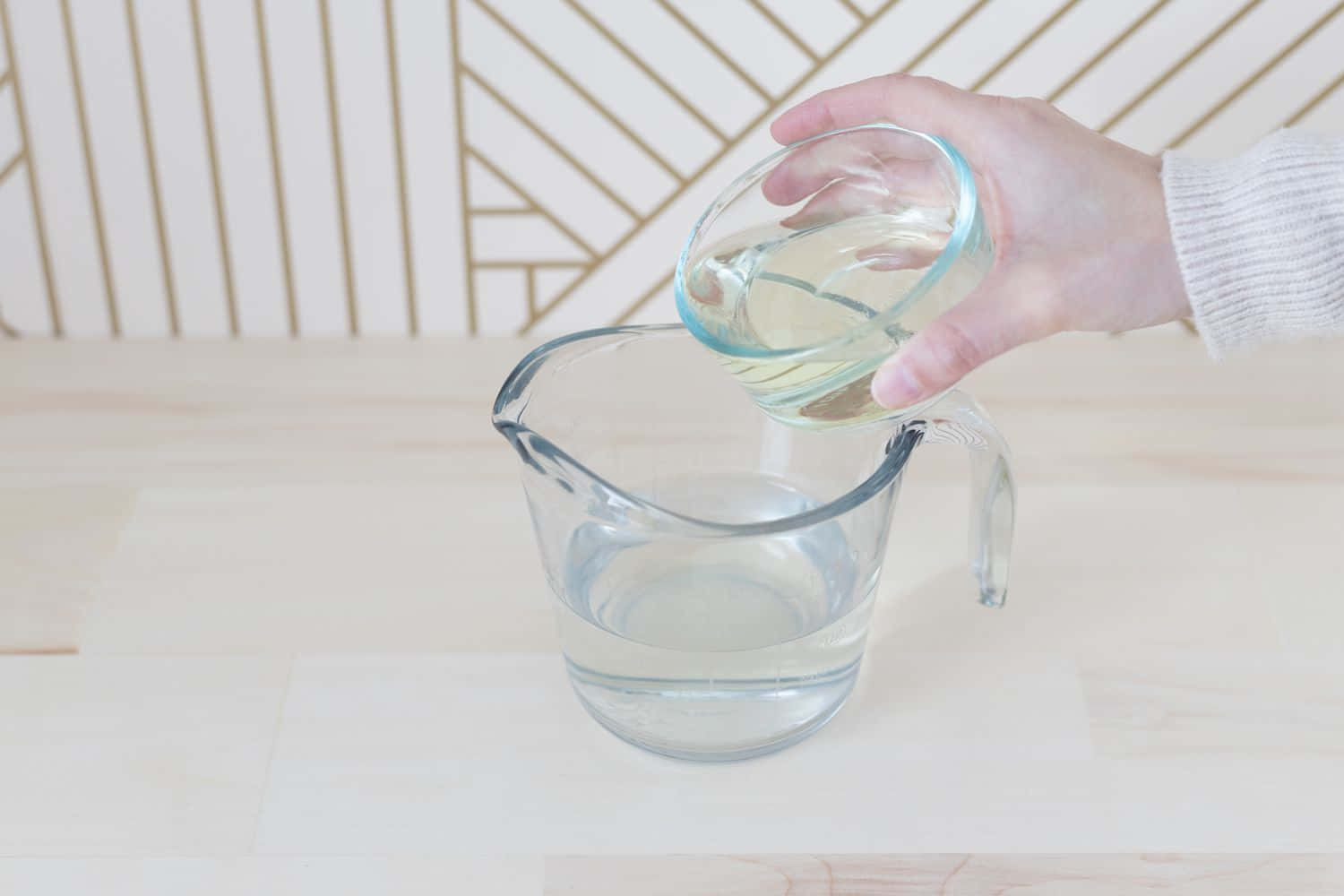 Cleaning 桌布 壁紙 — 一隻手將清澈液體從一個小玻璃碗倒入一個透明量杯，放在淺色木質表面上。