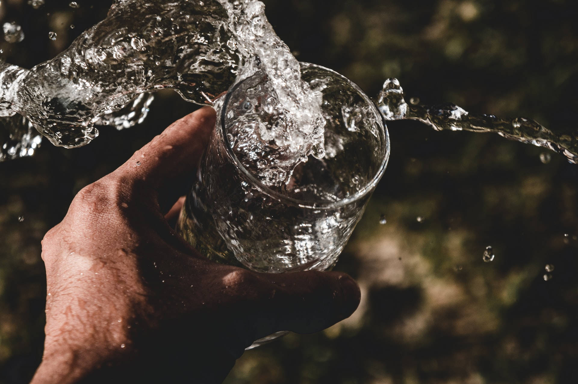 Moving Water 桌布 壁紙 — 一隻手將清澈的水倒入玻璃杯中，捕捉到動態的水花和水滴，背景是模糊的自然景色。