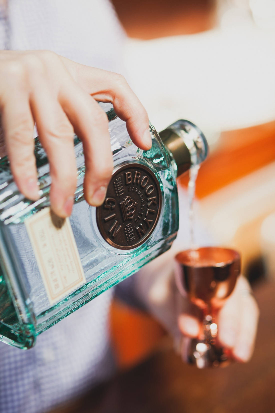 alcohol 桌布 壁紙 — 一隻手將清澈的琴酒從青綠色瓶子倒入銅製量酒器，背景溫暖模糊。