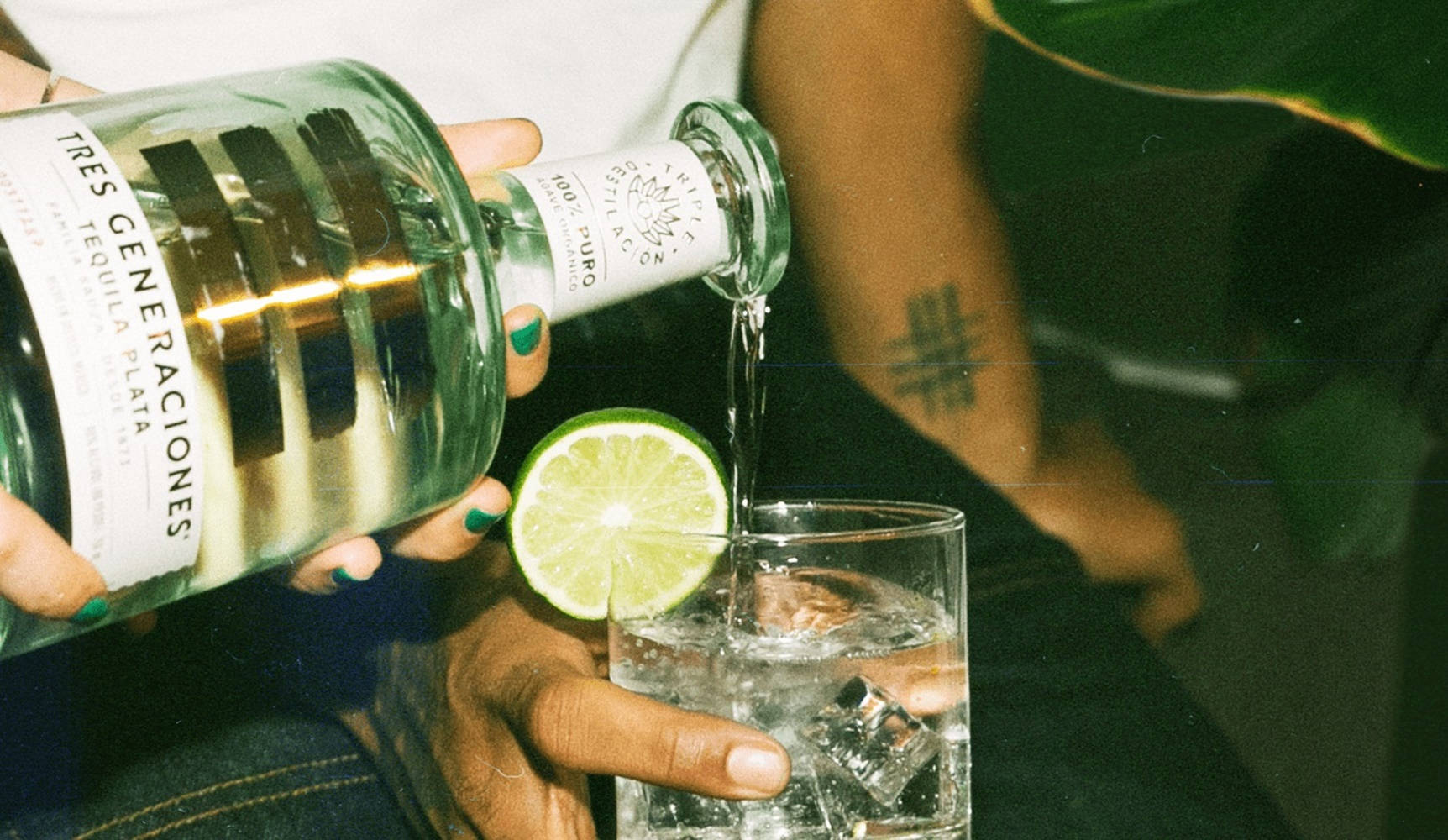 alcohol 桌布 壁紙 — 一隻手將清澈的龍舌蘭酒倒在冰塊上，倒入旁邊放有萊姆角的玻璃杯中，環境光線溫暖。