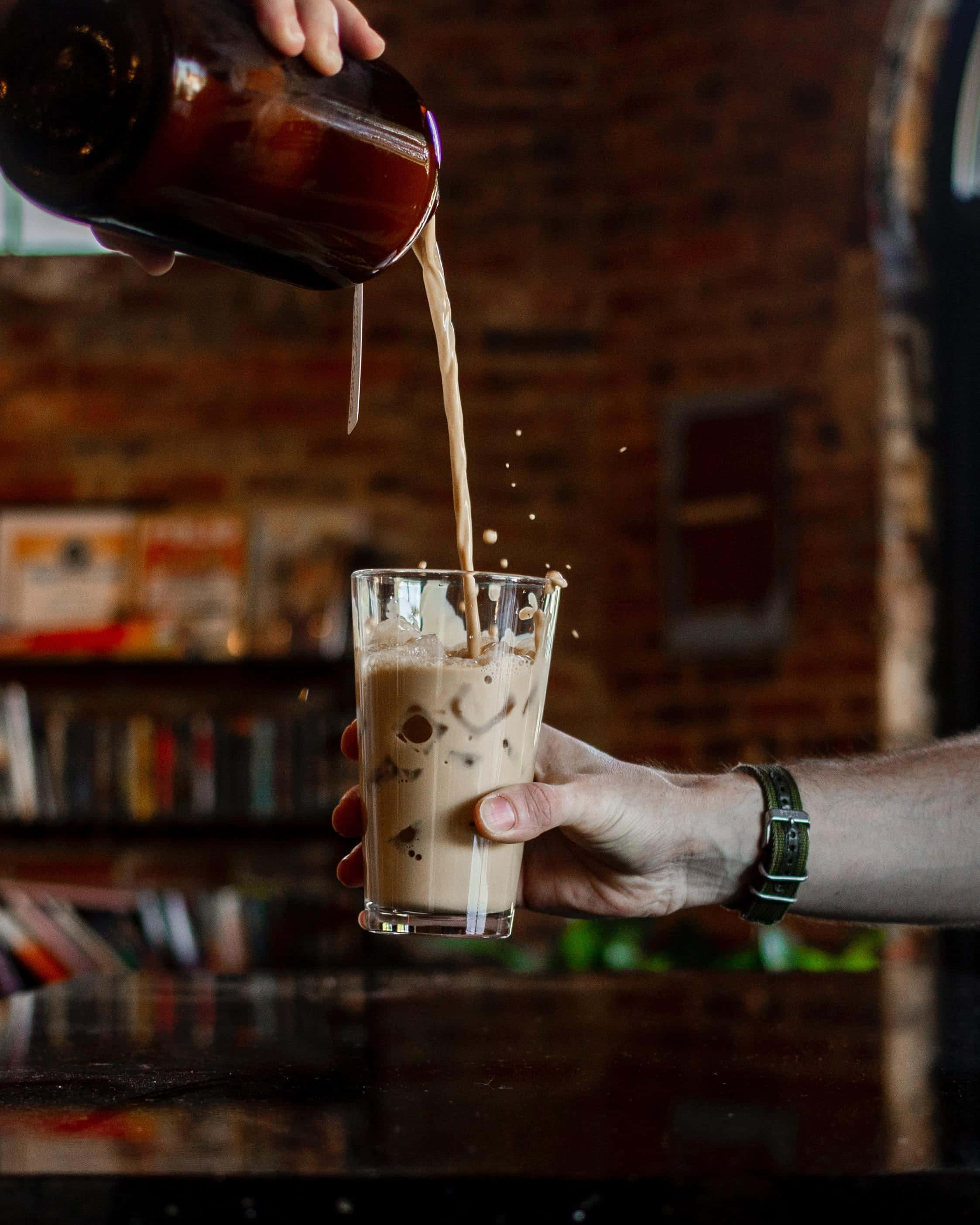 Iced Coffee 桌布 壁紙 — 一隻手將濃郁的咖啡倒在冰上，倒入一個靠在溫暖磚牆背景上的玻璃杯中。