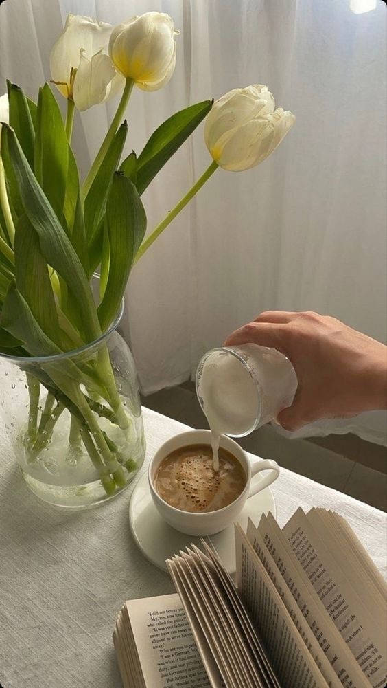 Coffee Aesthetic 桌布 壁紙 — 一隻手將牛奶倒入咖啡杯中，旁邊有白色鬱金香和一本打開的書，放在陽光照射的桌子上。