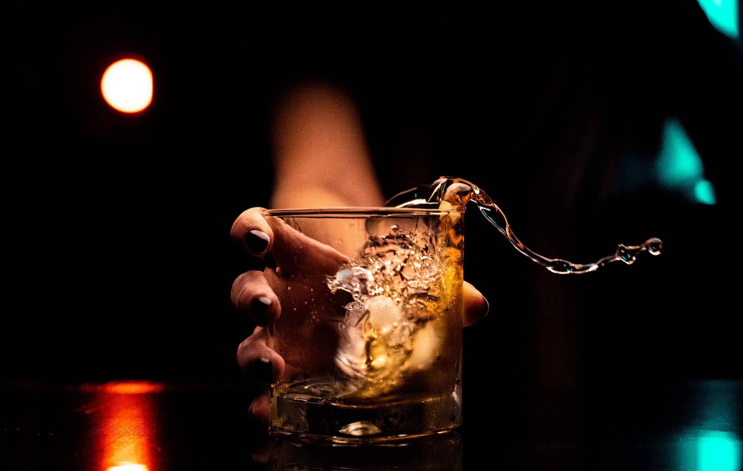 alcohol 桌布 壁紙 — 一隻手將琥珀色液體倒入裝有冰塊的玻璃杯中，背景昏暗，由溫暖與冷色調的散景燈光點亮。