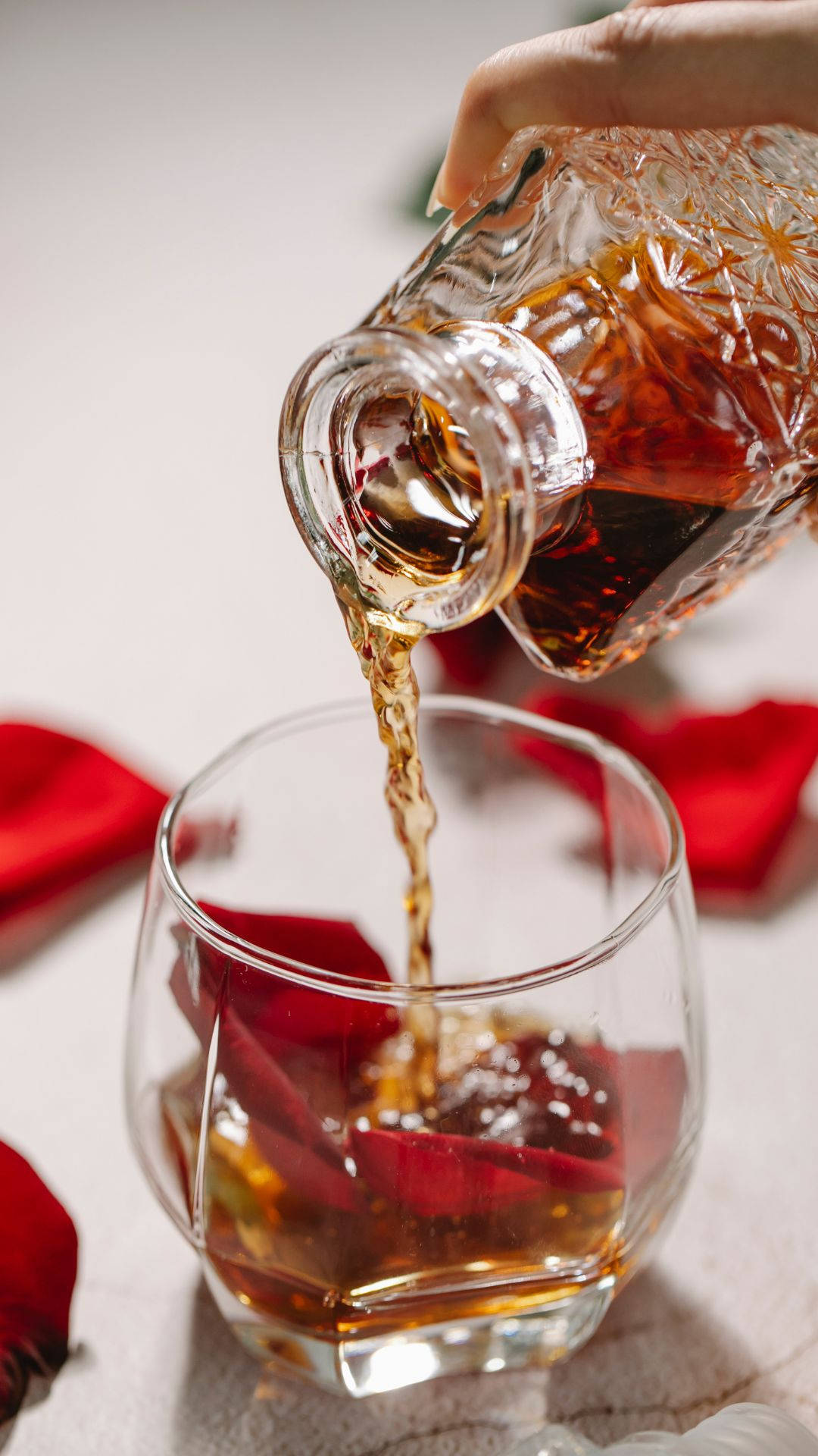alcohol 桌布 壁紙 — 一隻手將琥珀色液體從水晶醒酒器倒入一個飾有紅玫瑰花瓣的玻璃杯中，置於柔軟的白色表面上。