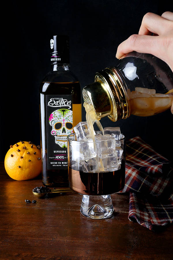cocktail 桌布 壁紙 — 一隻手將琥珀色酒液倒在冰塊上，倒入玻璃杯中，旁邊放著骷髏標籤的酒瓶和一個香料橙，置於深色木桌上。