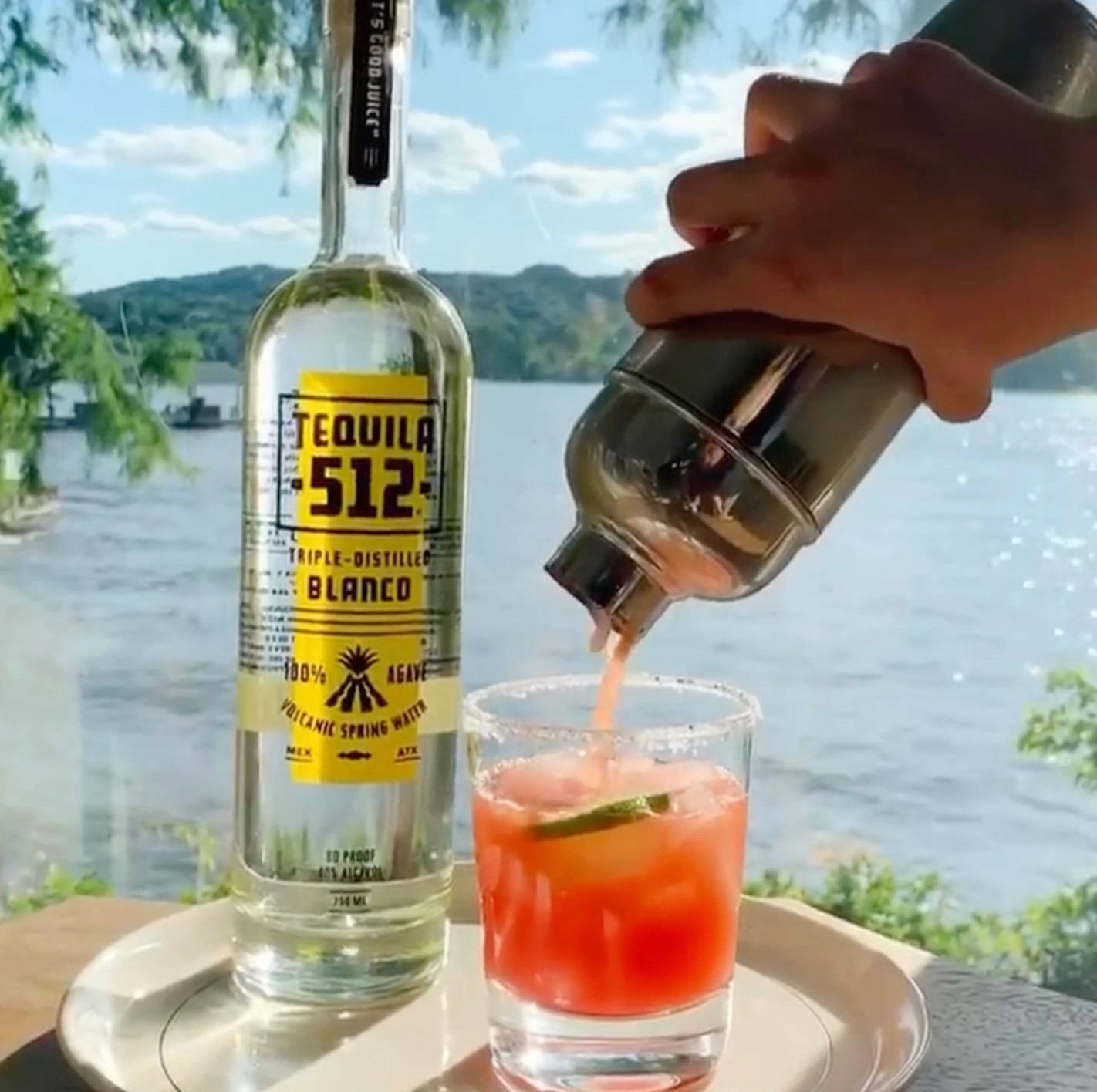 drinks 桌布 壁紙 — 一隻手將調酒器中的鮮豔粉紅雞尾酒倒入鹽邊玻璃杯，旁邊放著一瓶Tequila 512，背景是明亮的湖邊景色。