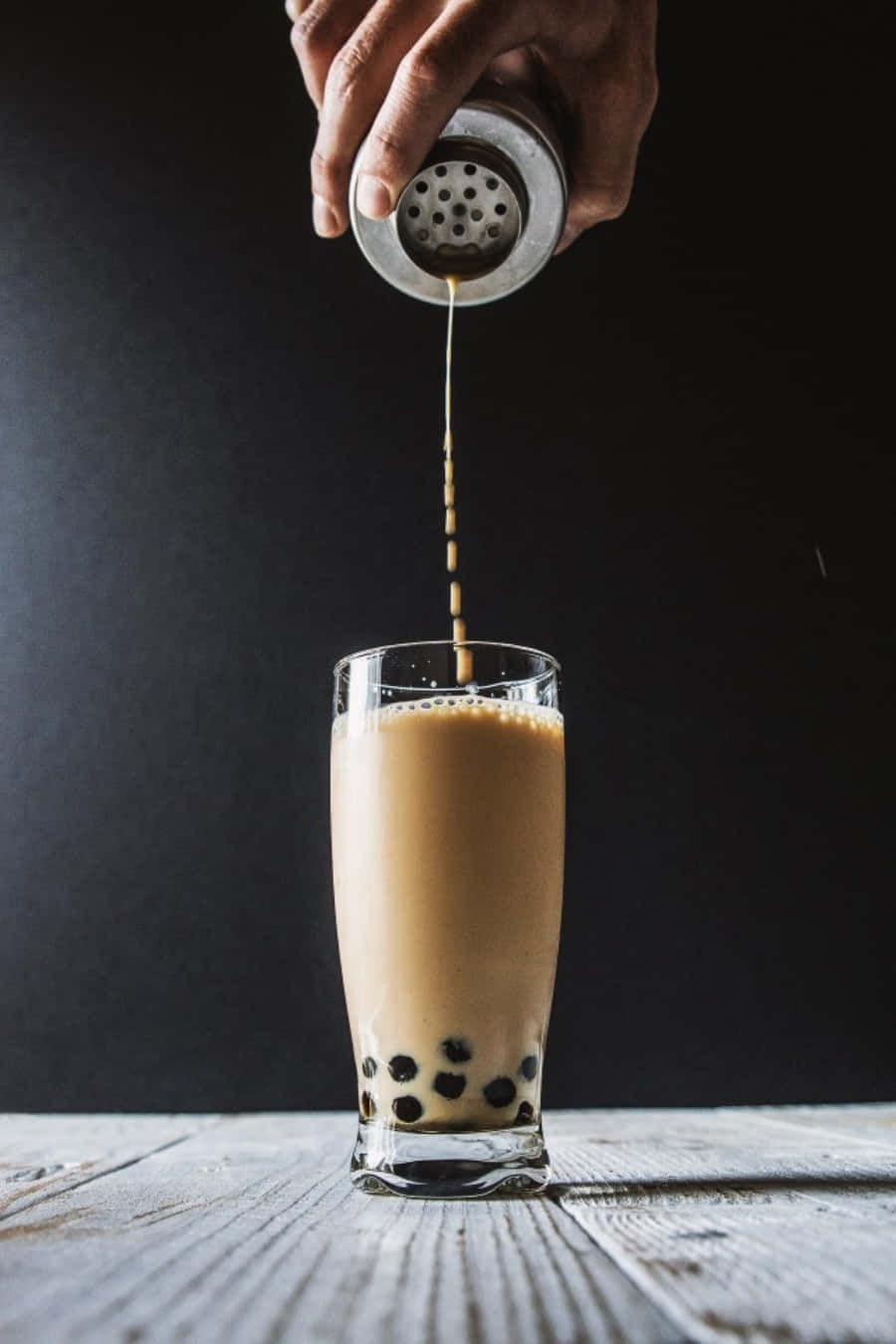 Boba Tea 桌布 壁紙 — 一隻手將金屬搖杯中的奶油泡沫奶茶倒入裝有木薯珍珠的玻璃杯中，放在質樸的木質表面上。