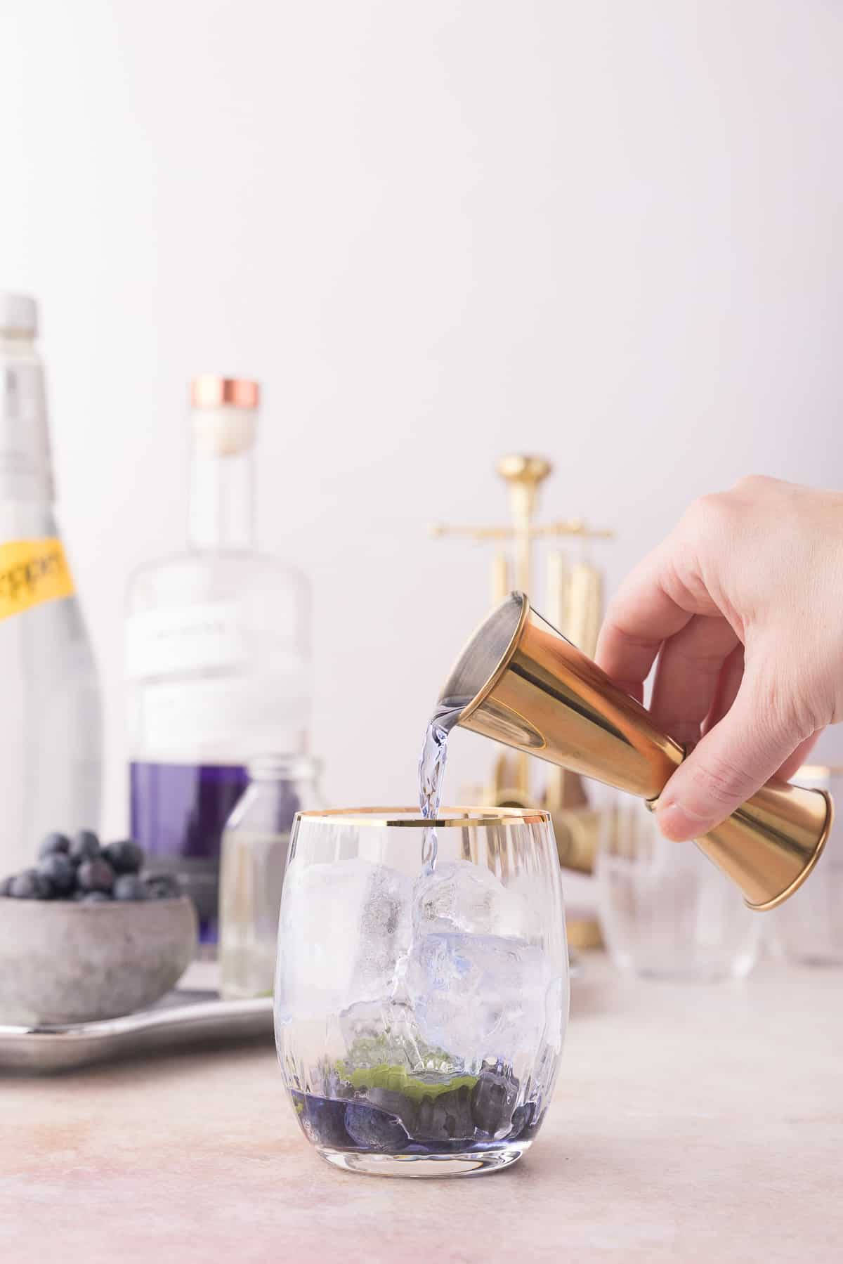 cocktail 桌布 壁紙 — 一隻手將金色量酒器中的清澈液體倒入裝滿冰塊和藍莓的玻璃杯中。