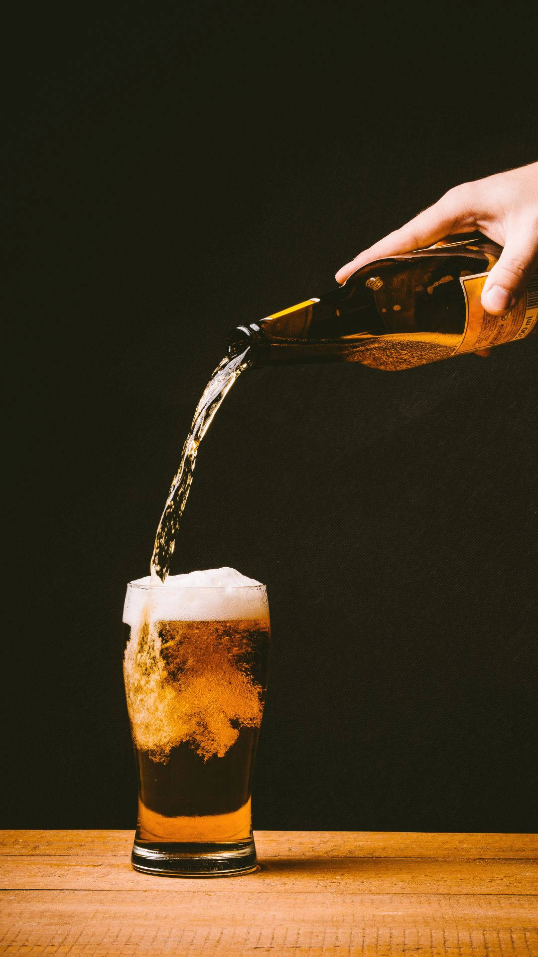 alcohol 桌布 壁紙 — 一隻手將金黃色啤酒倒入木桌上的玻璃杯中，背景為深色。