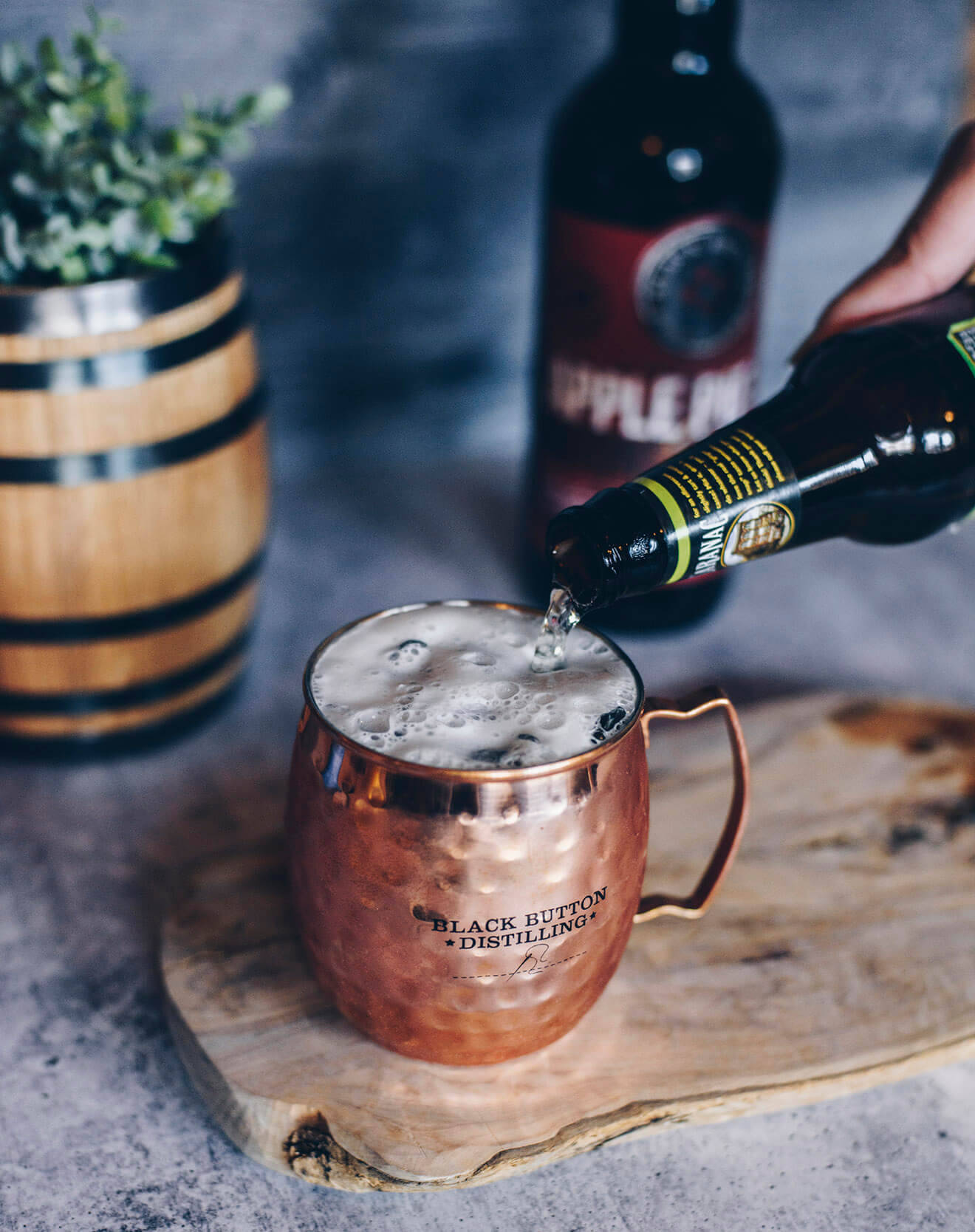 pour one out 桌布 壁紙 — 一隻手將黑啤酒倒入一個印有 black button distilling 標誌的錘紋銅杯，放在質樸的木板上。