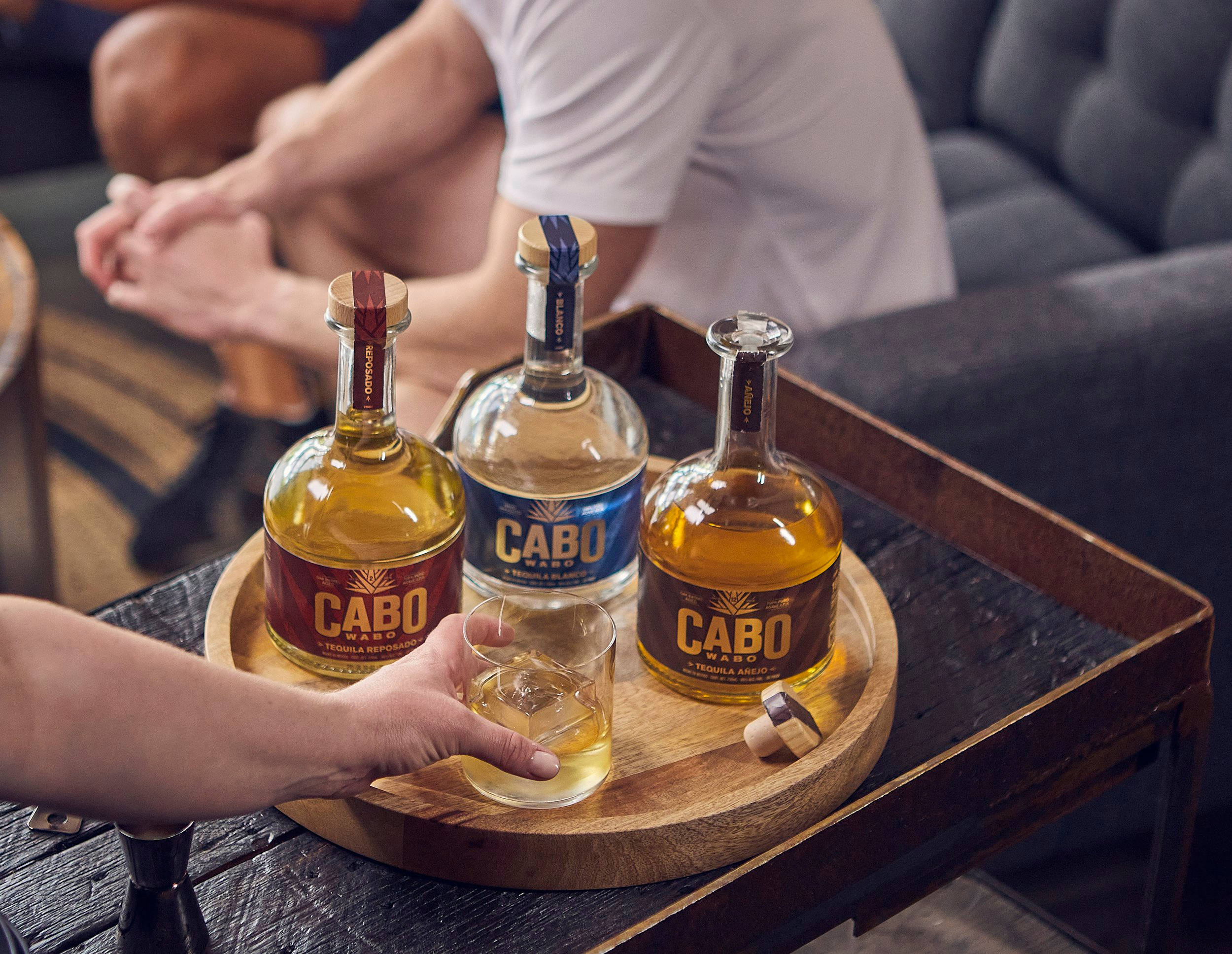 Drink 桌布 壁紙 — 一隻手將龍舌蘭酒從酒瓶倒到一個木托盤上，托盤上有三瓶Cabo Wabo酒和一個加冰的玻璃杯。