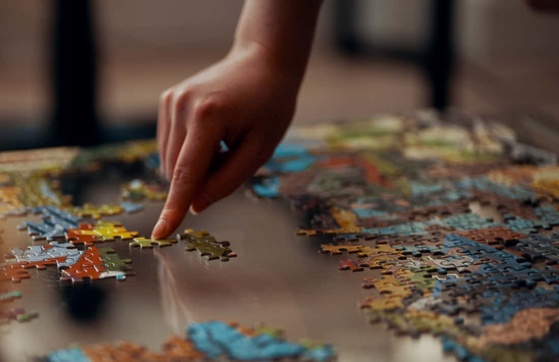 Puzzle 桌布 壁紙 — 一隻手小心地將一塊拼圖放在反射桌上部分完成的彩色拼圖上的特寫。