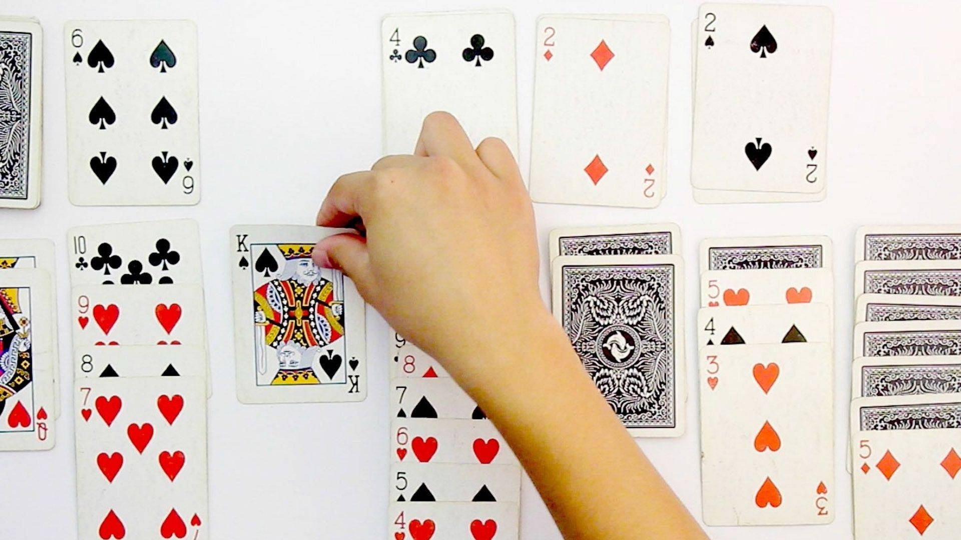 Solitaire 桌布 壁紙 — 一隻手小心地將一張黑桃K牌放在白色平面上散落的紙牌中。