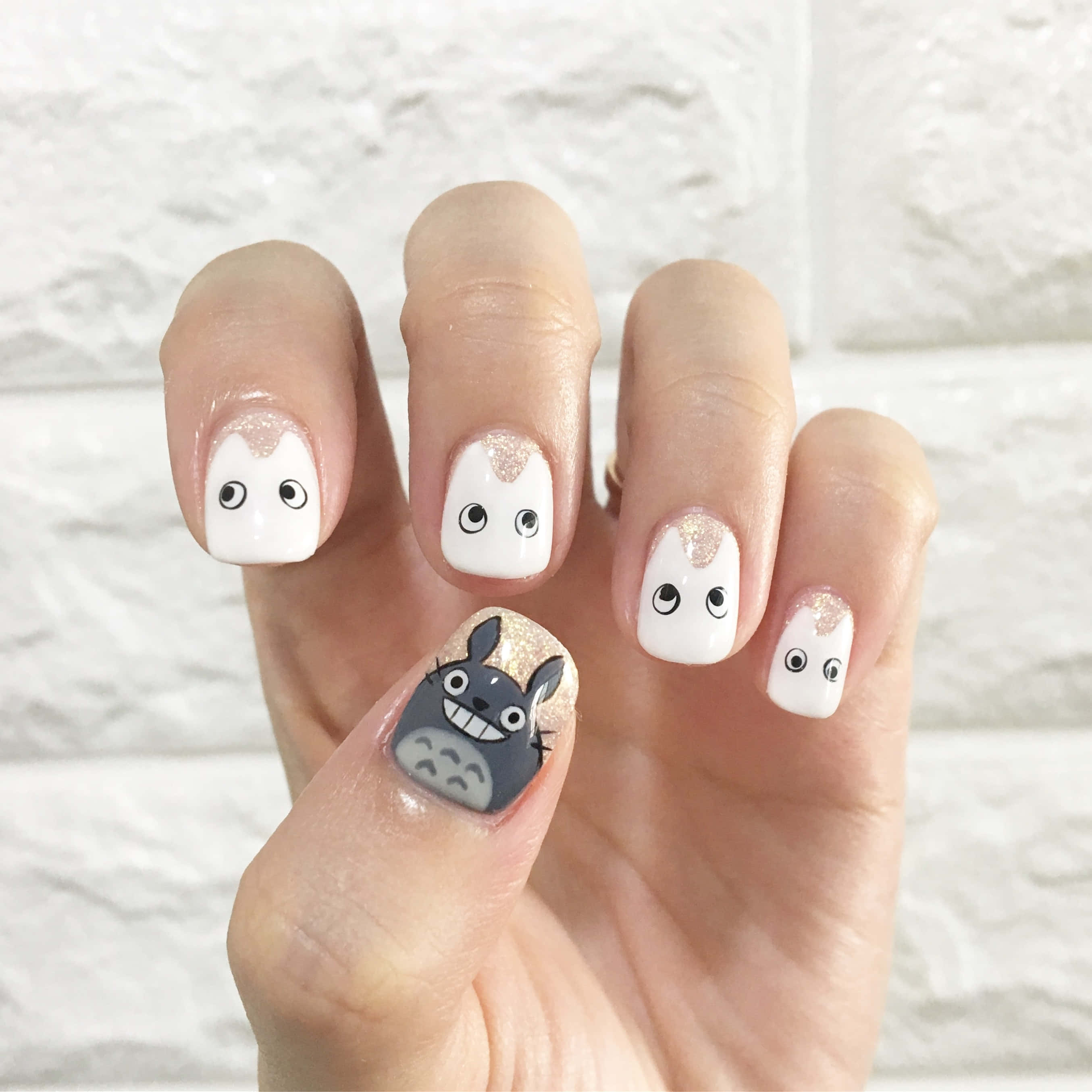 Nail 桌布 壁紙 — 一隻手展示可愛的龍貓主題美甲，採用閃亮白色和灰色設計，背景是帶紋理的白色磚牆。