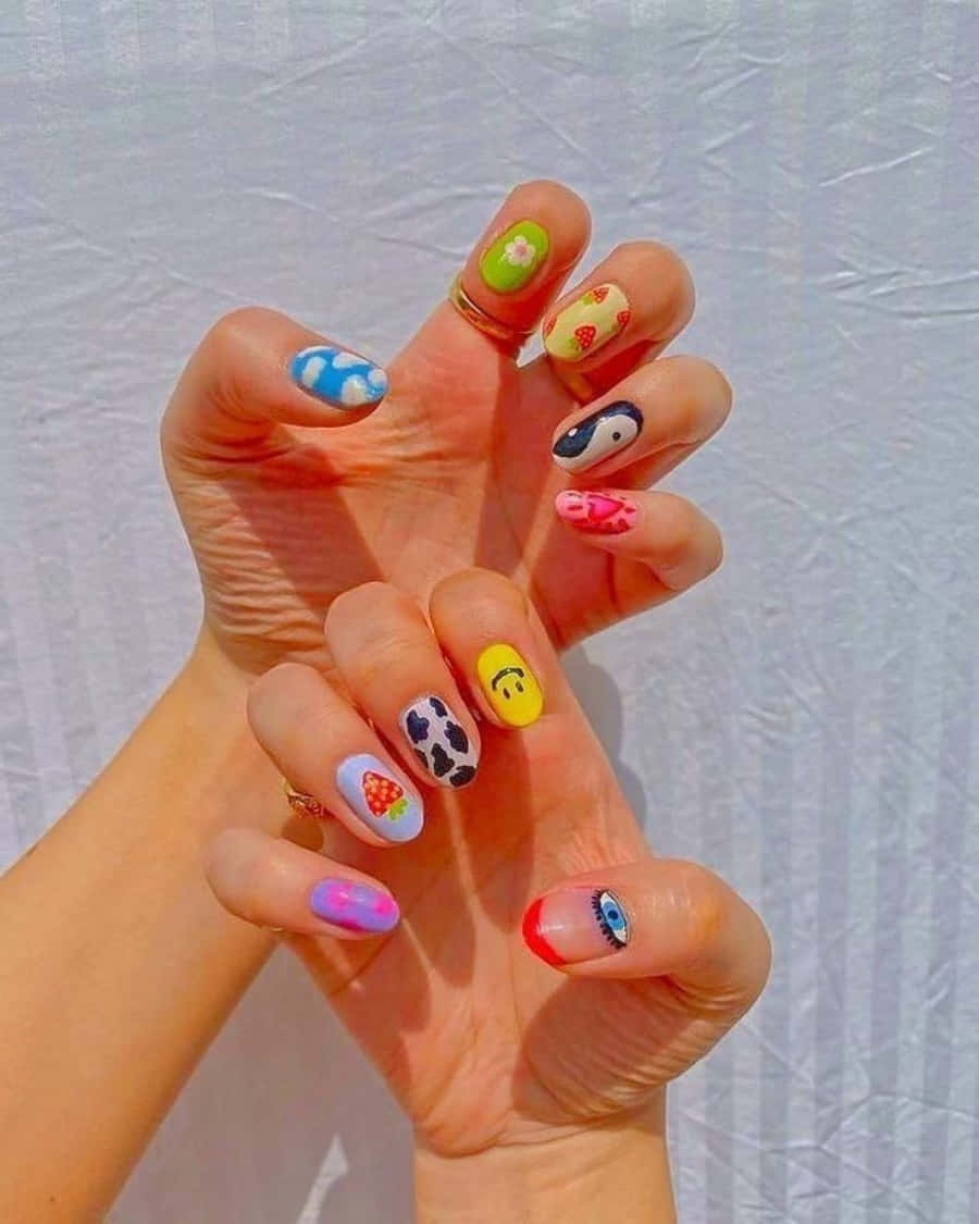 Nail 桌布 壁紙 — 一隻手展示色彩繽紛的卡通主題指甲藝術，圖案包括笑臉、草莓和雲朵，背景為皺巴巴的白色。