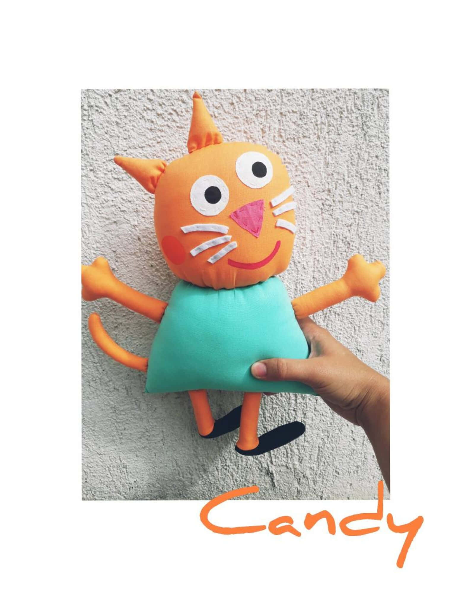 Candy Cat 桌布 壁紙 — 一隻手工製作的橙色貓咪絨毛玩具穿著青綠色連衣裙，被舉起靠在有紋理的灰色牆上。