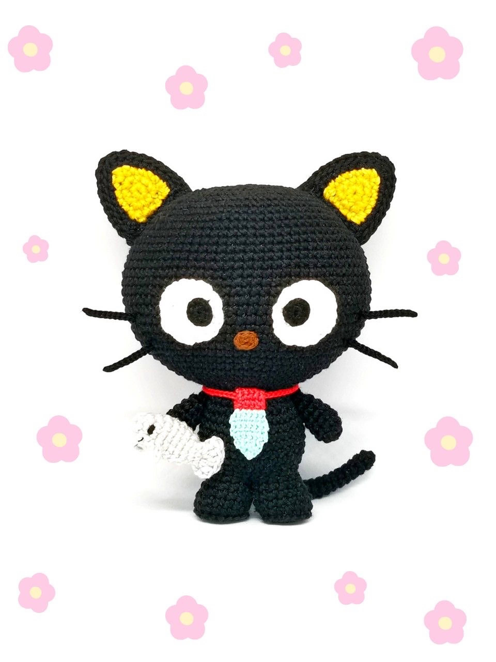 Chococat 桌布 壁紙 — 一隻手工製作的黑色鉤針貓咪，黃色耳朵和紅色蝴蝶結，抱著一條白色的魚，背景是粉紅色的花朵。