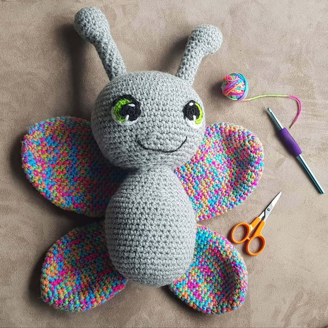 Crochet 桌布 壁紙 — 一隻手工鉤織的蝴蝶，灰色身體，彩虹翅膀，安放在米色表面上，旁邊有手工工具。