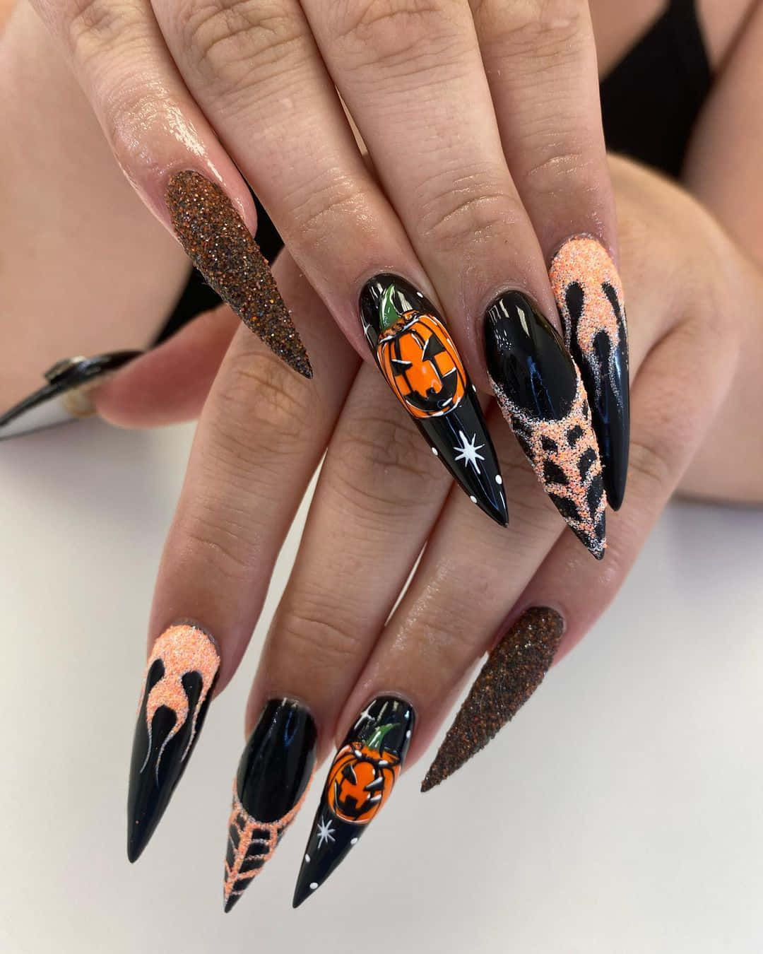 Halloween Nail Art 桌布 壁紙 — 一隻手帶有長尖形指甲，飾有黑色閃粉橙色南瓜燈和蜜桃豹紋萬聖節設計。