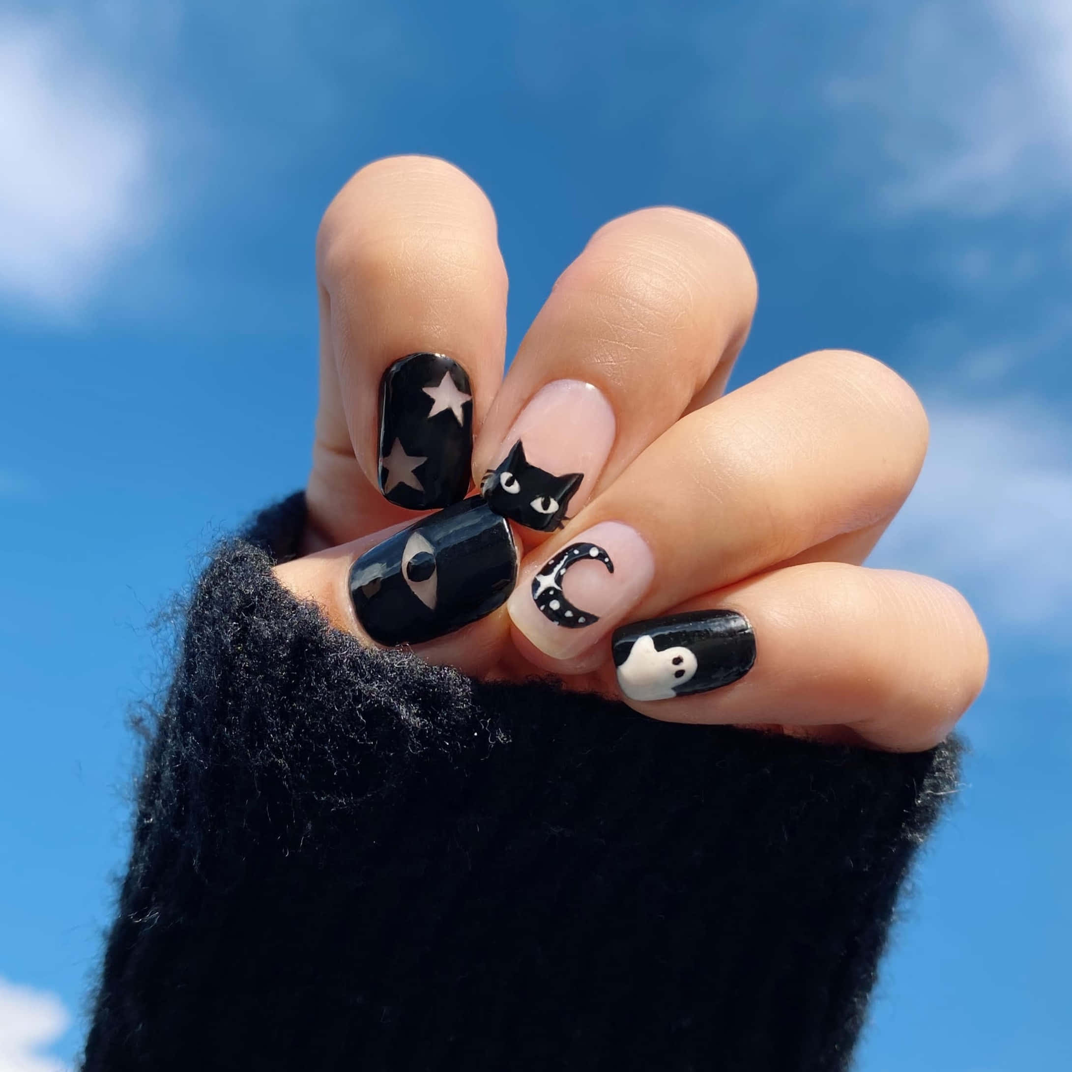 Halloween Nail Art 桌布 壁紙 — 一隻手帶有黑白女巫主題的美甲，背景是明亮的藍天和蓬鬆的雲朵。